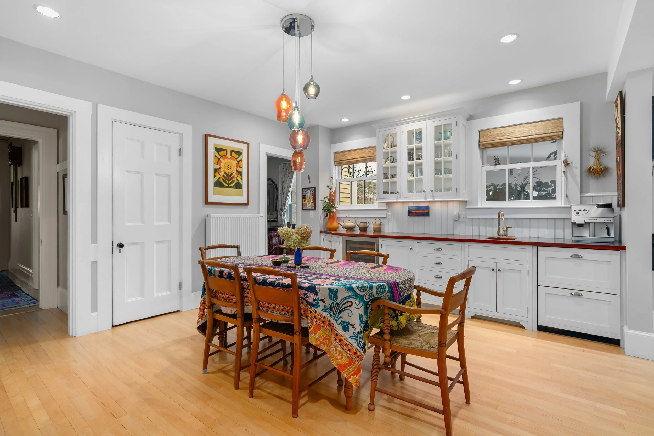 Bright kitchen with white cabinets, a wooden dining table with a colorful tablecloth, and hanging pendant lights with glass shades.