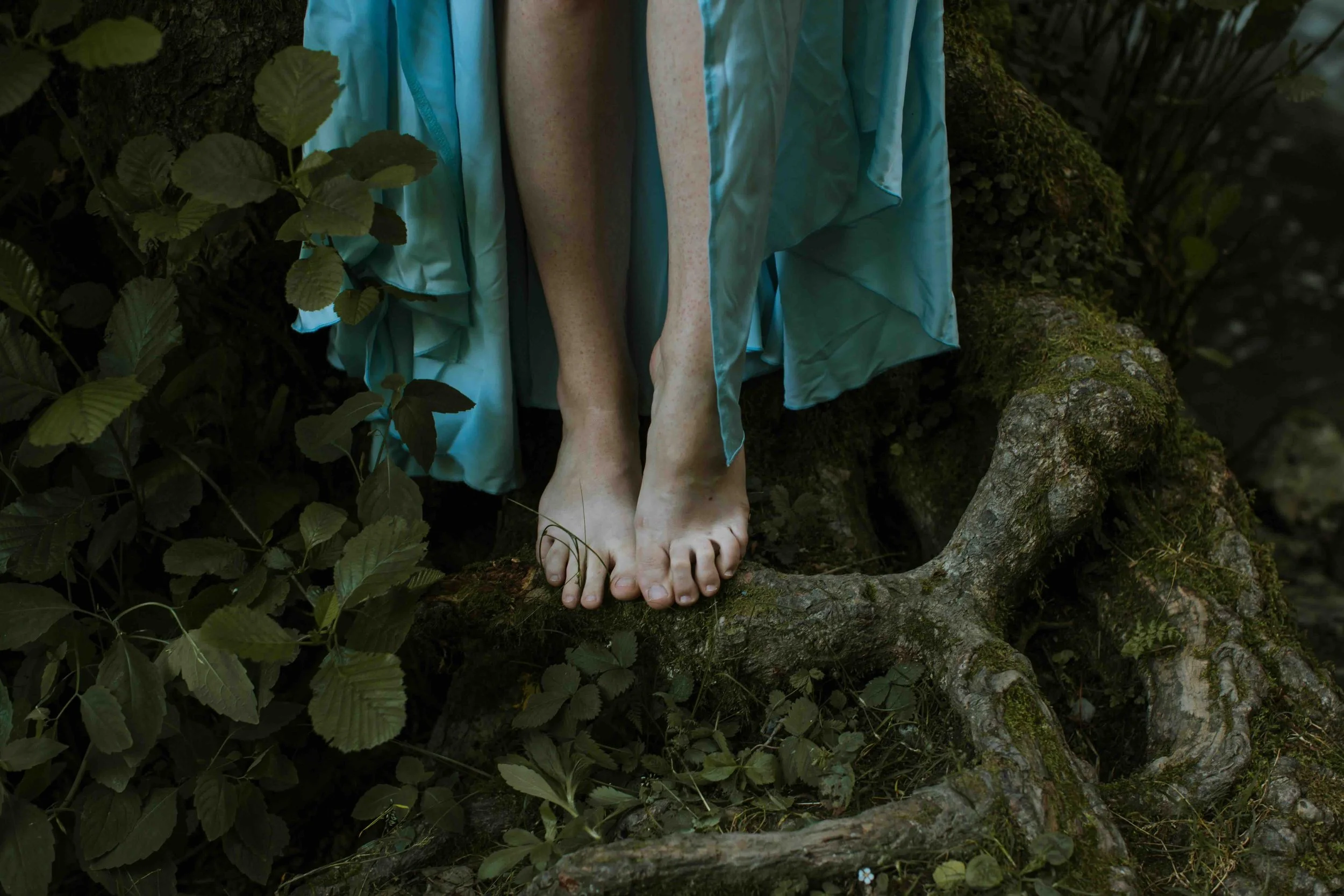 Close-up of a person's feet with light skin tone, standing on a moss-covered tree root in a forest, wearing a flowing, light blue garment.