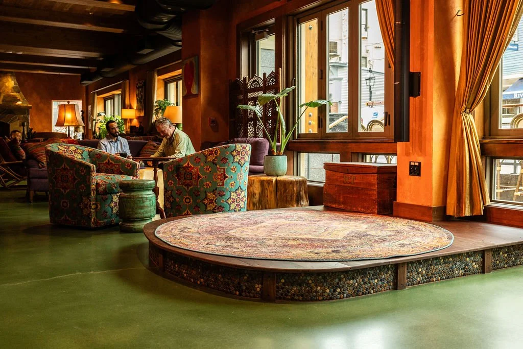 Cozy lounge with patterned armchairs, a circular rug, potted plant, large windows with yellow curtains, and a few people sitting and talking.