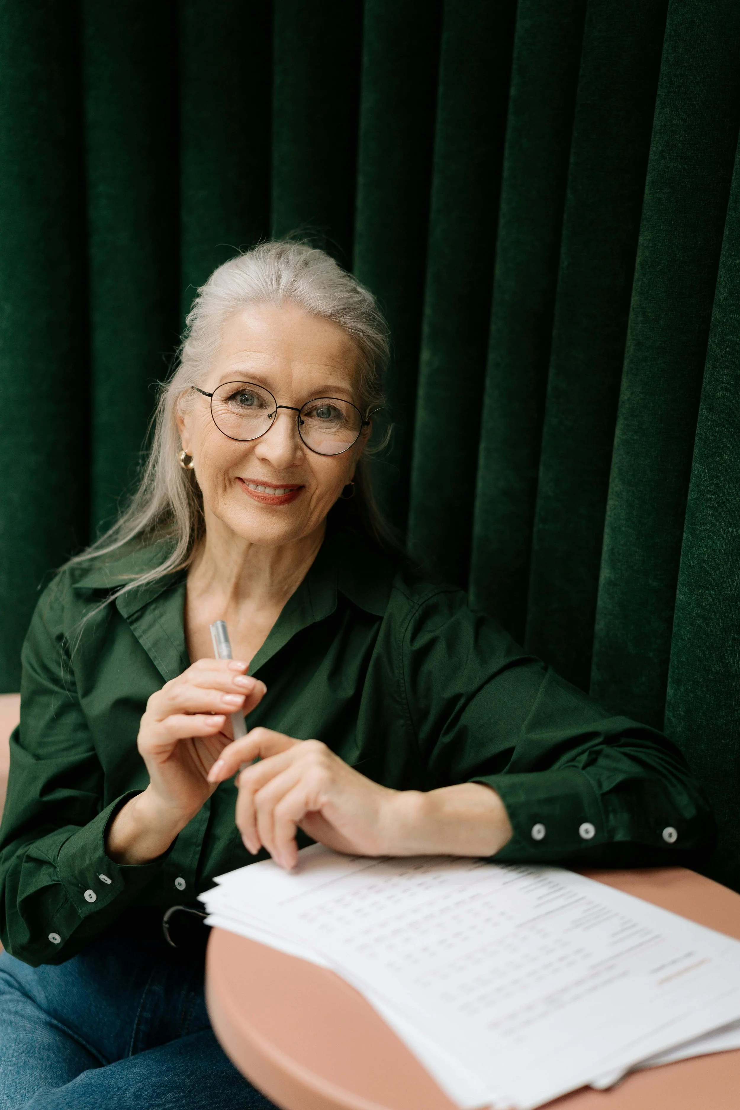 An elderly woman with glasses, gray hair, and earrings, smiling and holding a pen, sitting at a table with papers, against a background of dark green curtains.