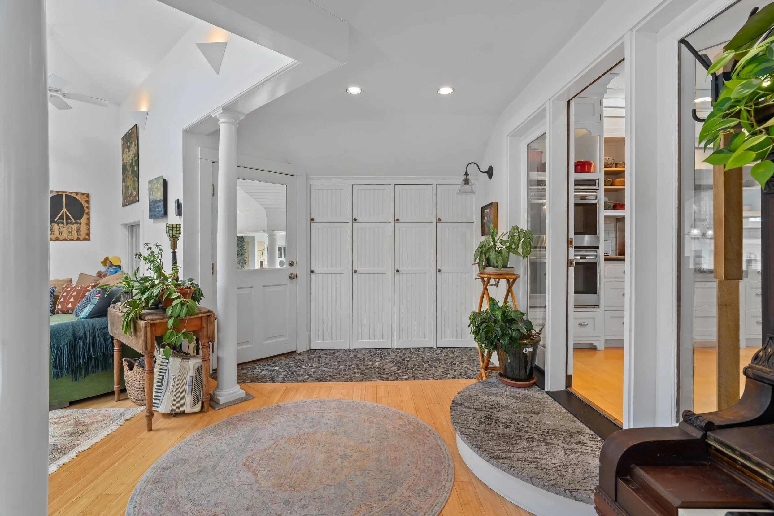 Interior view of a house entryway with white walls, a white door, closet doors, and various potted plants, with a glimpse of the living room on the left and the kitchen through a doorway on the right