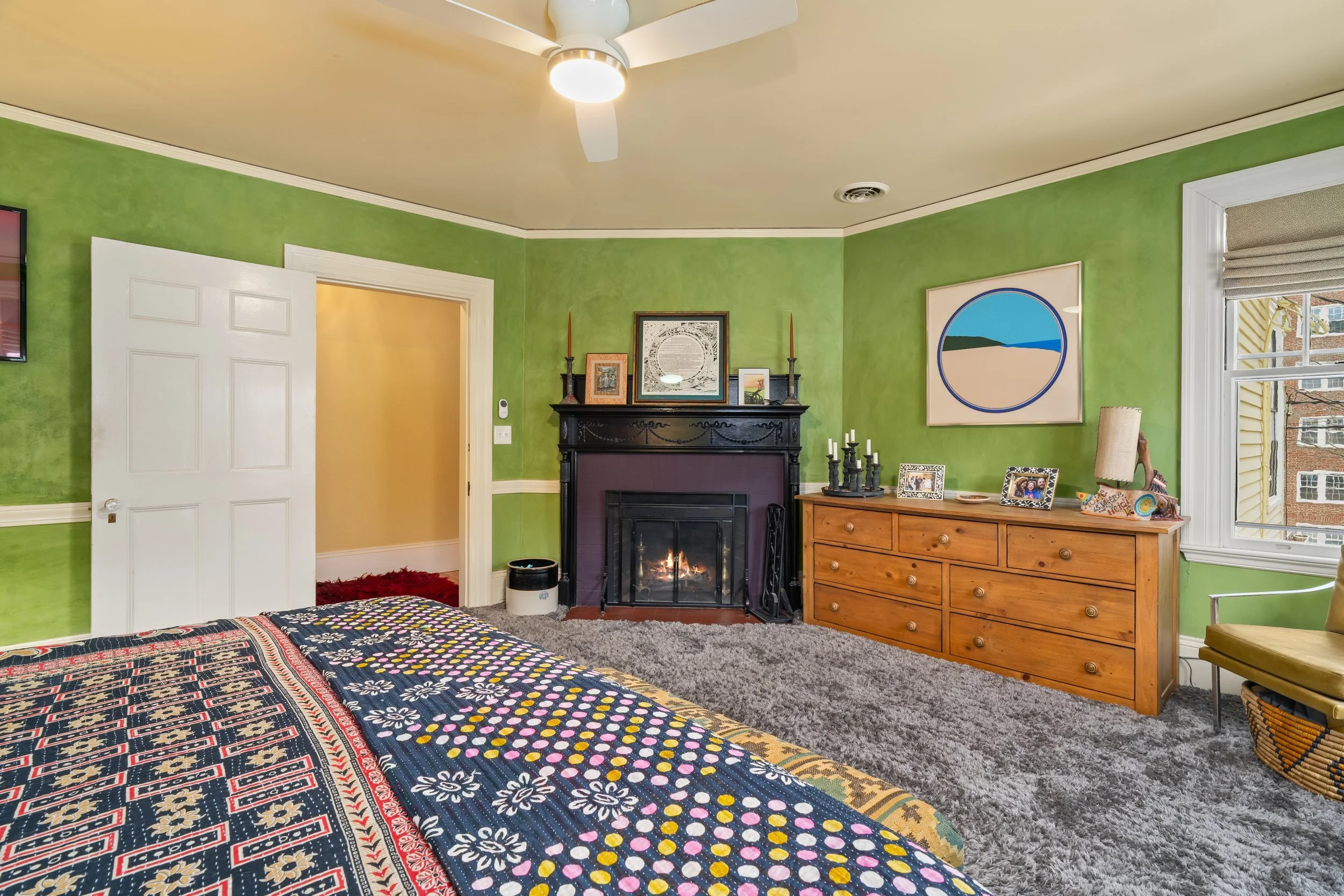 A bedroom with green walls, a fireplace with black and purple accents, a wooden dresser, a window with blinds, a yellow chair, and a ceiling fan with a light. The bed has a patterned quilt, and there are various decorative items and framed photos on