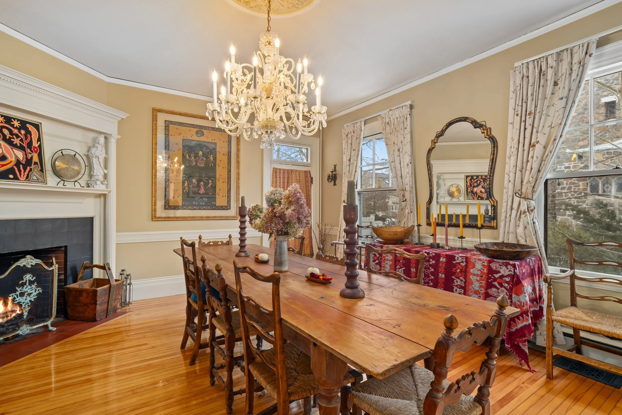 A dining room with a wooden table, seven chairs, a chandelier, a fireplace, and vintage decor. Windows with floral curtains are visible.