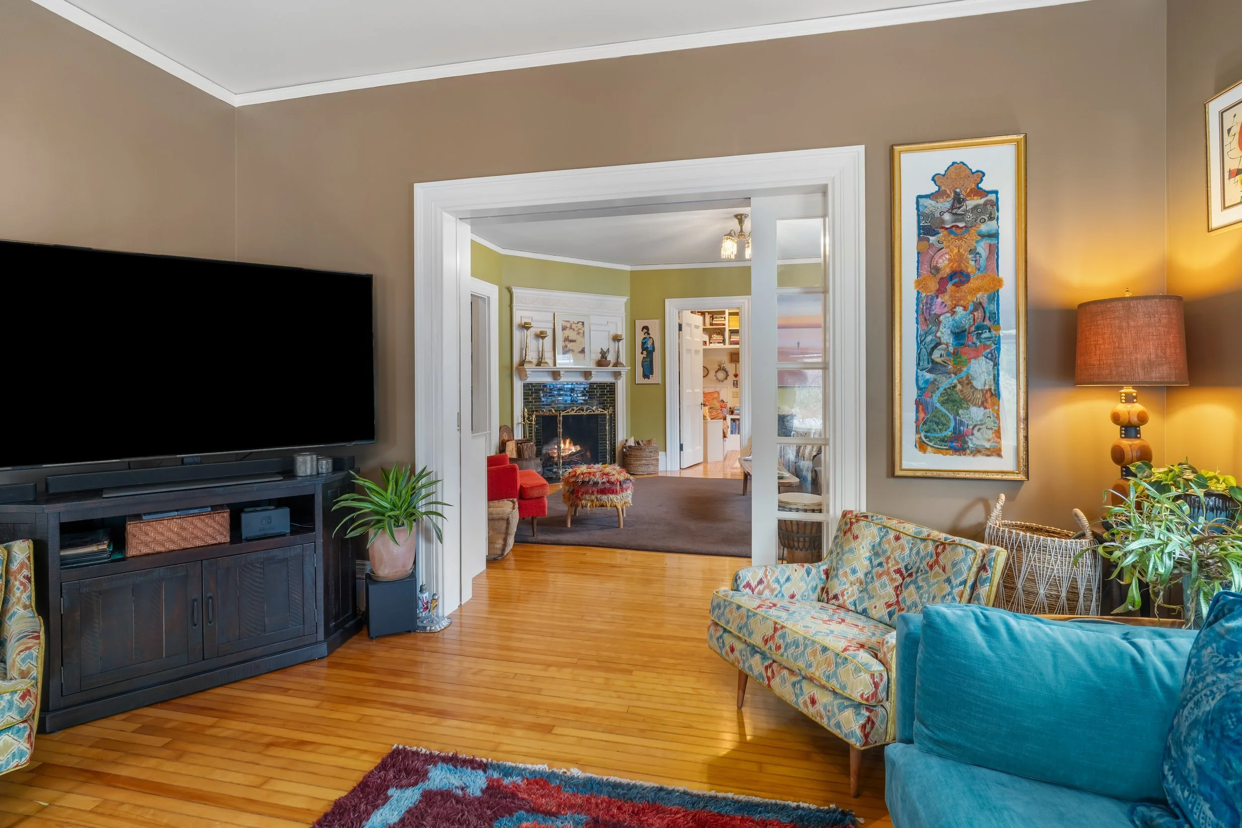 Living room with beige walls, hardwood floors, a large flat-screen TV on a black stand, colorful patterned couches, a side table with a plant, wall art, and a lamp, connected to a cozy den with a fireplace and chairs.
