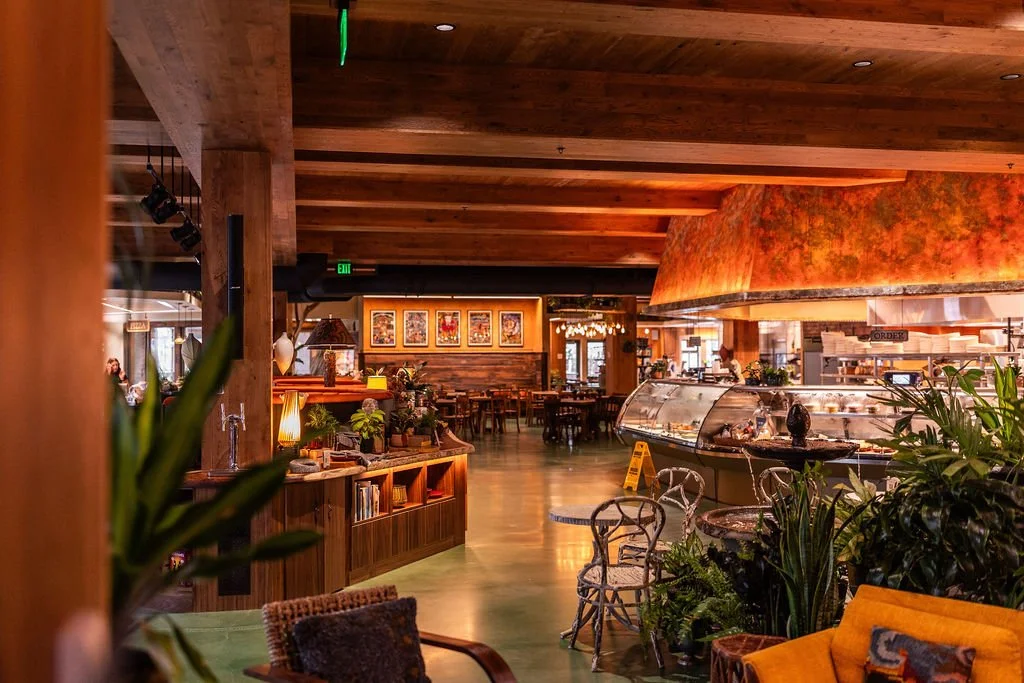 Interior of a cozy restaurant with wooden decor, artwork on the walls, a glass display case, and various seating options including chairs and sofas surrounded by plants.