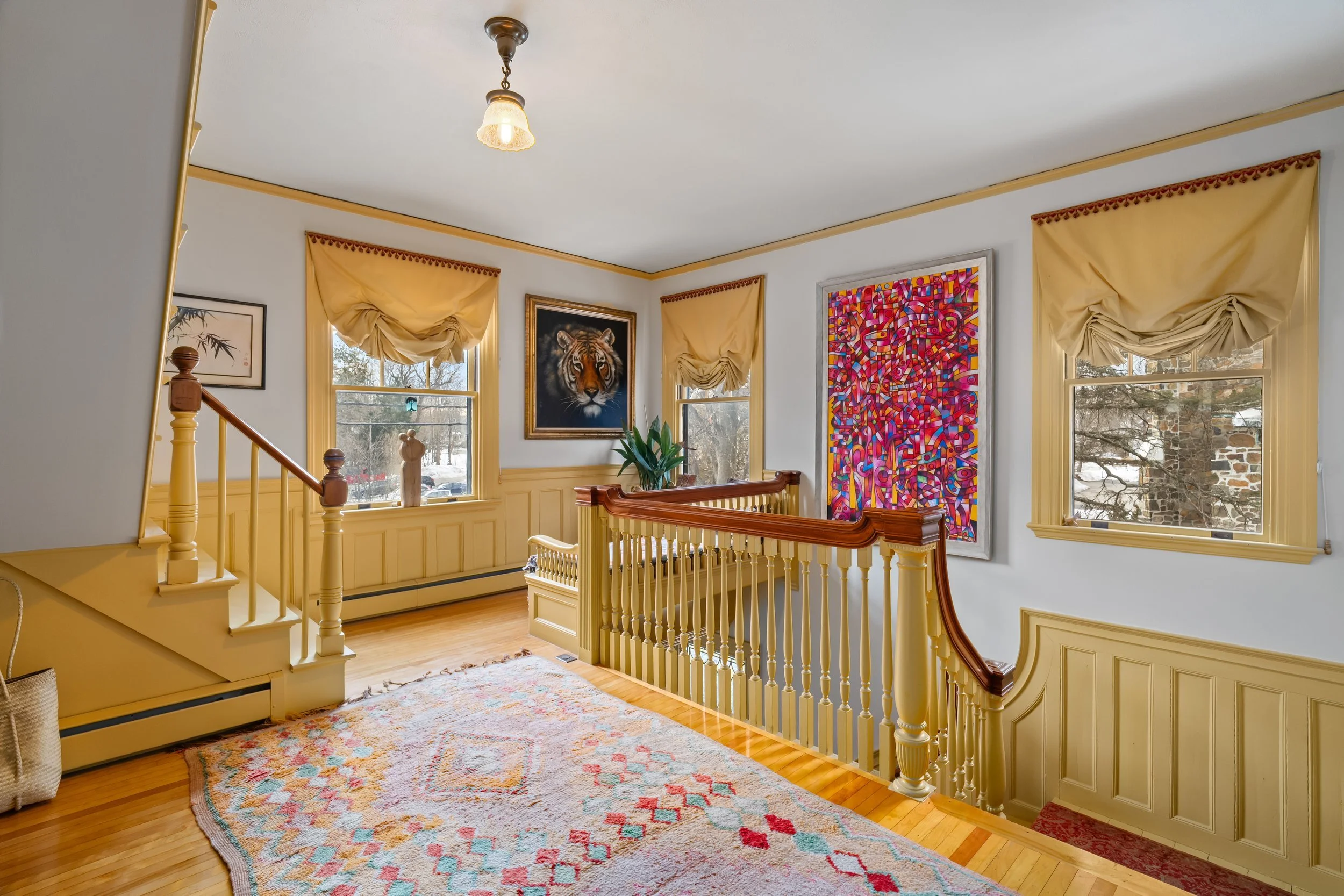 A bright living room with three windows featuring yellow curtains, a wooden staircase with yellow balusters, a multicolored abstract painting on the wall, a portrait of a lion, a potted plant, and a light-colored area rug on a wooden floor.