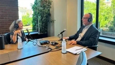 Two people sitting across from each other at a table, wearing headphones and speaking into microphones, likely recording a podcast or radio interview, with large windows and greenery outside.