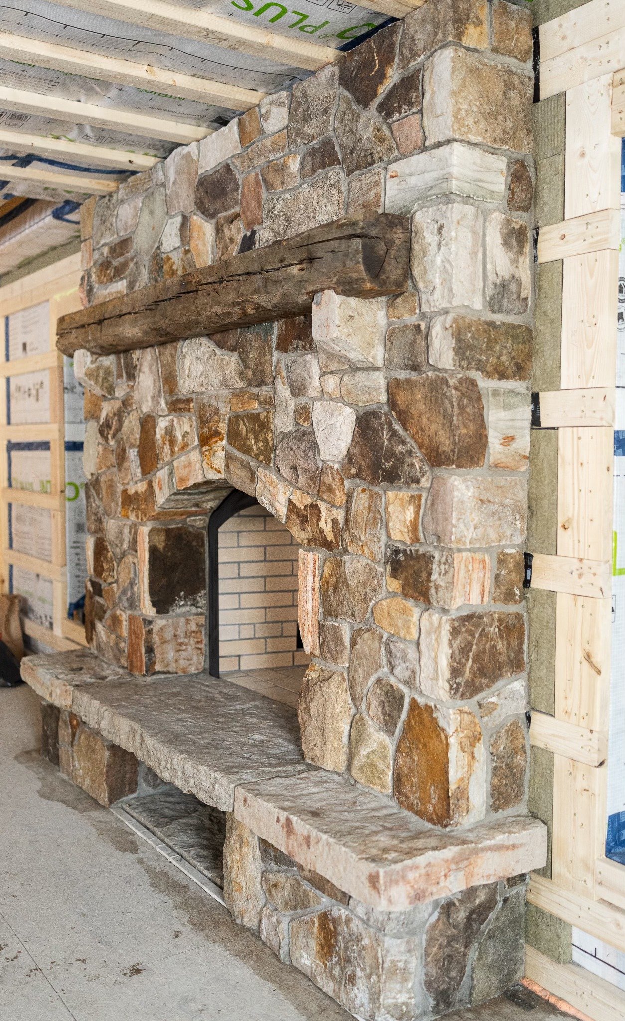 A stone fireplace with a wooden mantel under construction in a building with exposed framing and insulation.