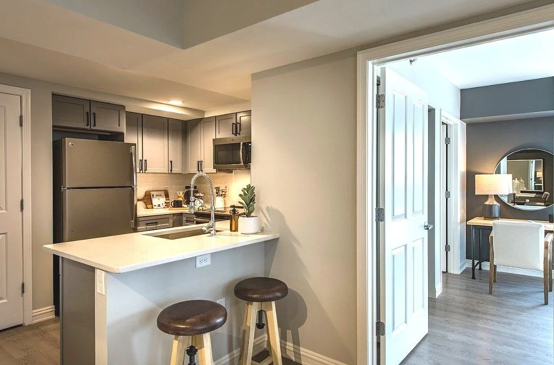 Modern kitchen with gray cabinets, stainless steel refrigerator, black microwave, white countertop, and small bar stools. Adjacent room with white chairs and a lamp, visible through open double doors.