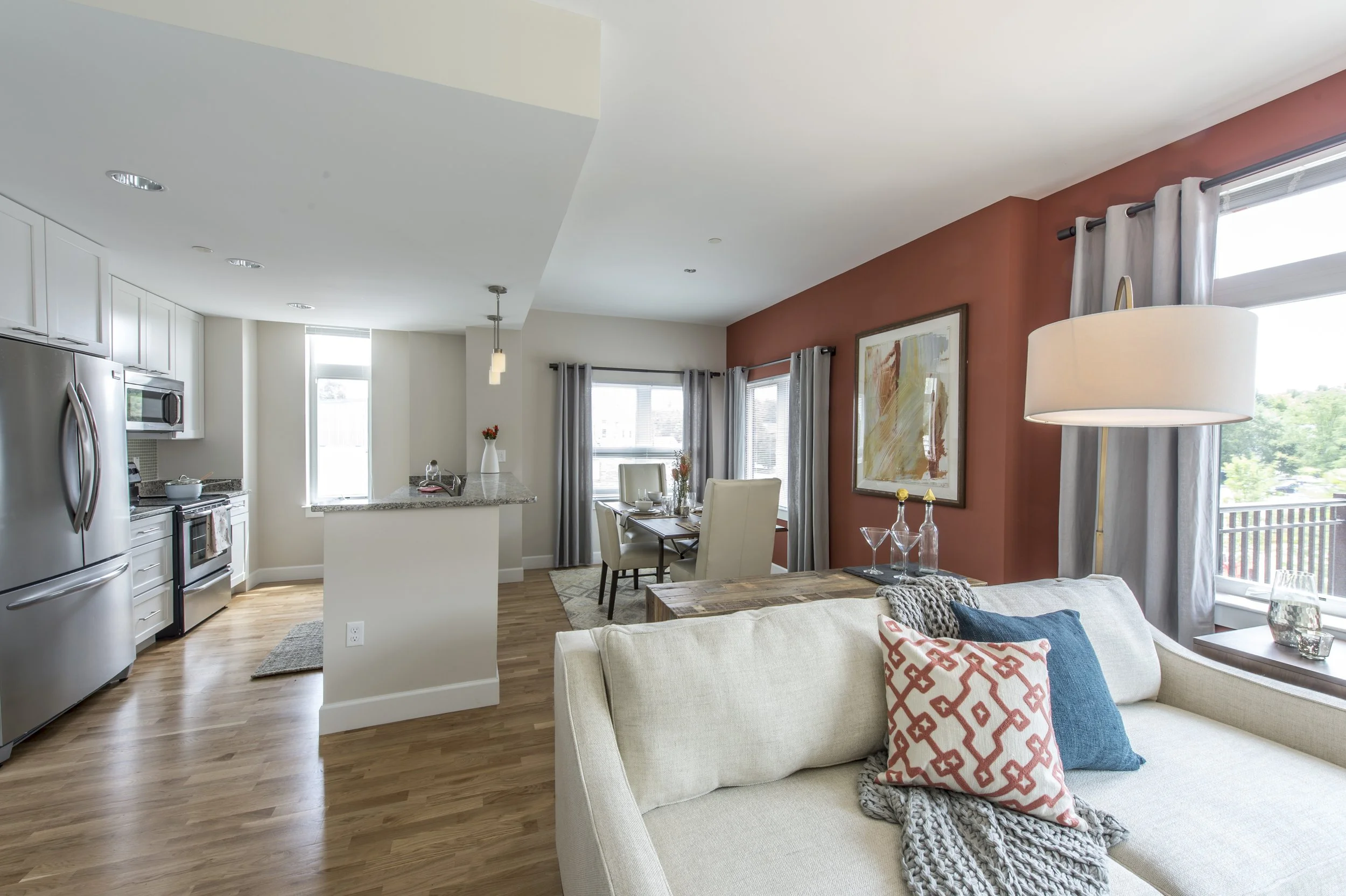 Open-concept living room with beige sofa, decorative pillows, wooden coffee table, large windows with curtains, and a dining area with a table and chairs. Kitchen in the background with stainless steel appliances and white cabinetry.