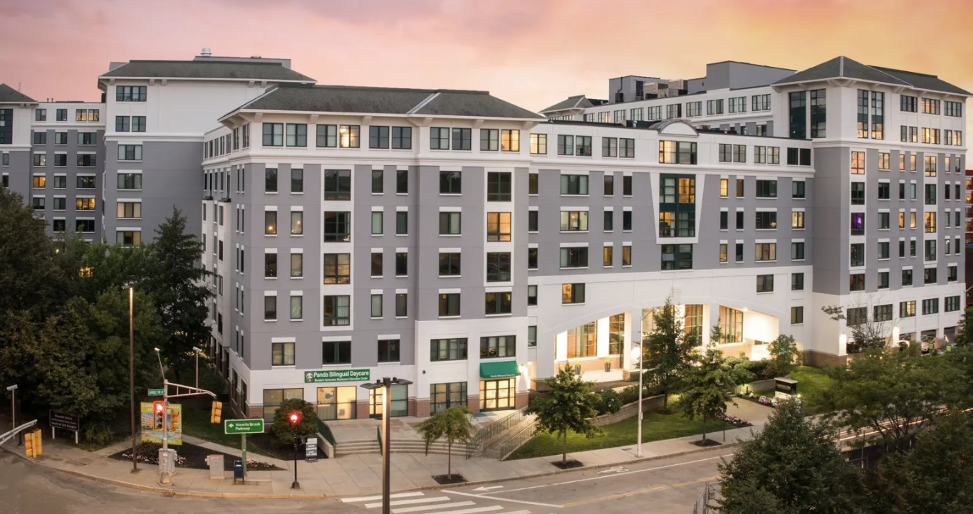 A multi-story modern apartment building at dusk with illuminated windows, surrounding trees, sidewalk, streetlights, and a pedestrian crossing.