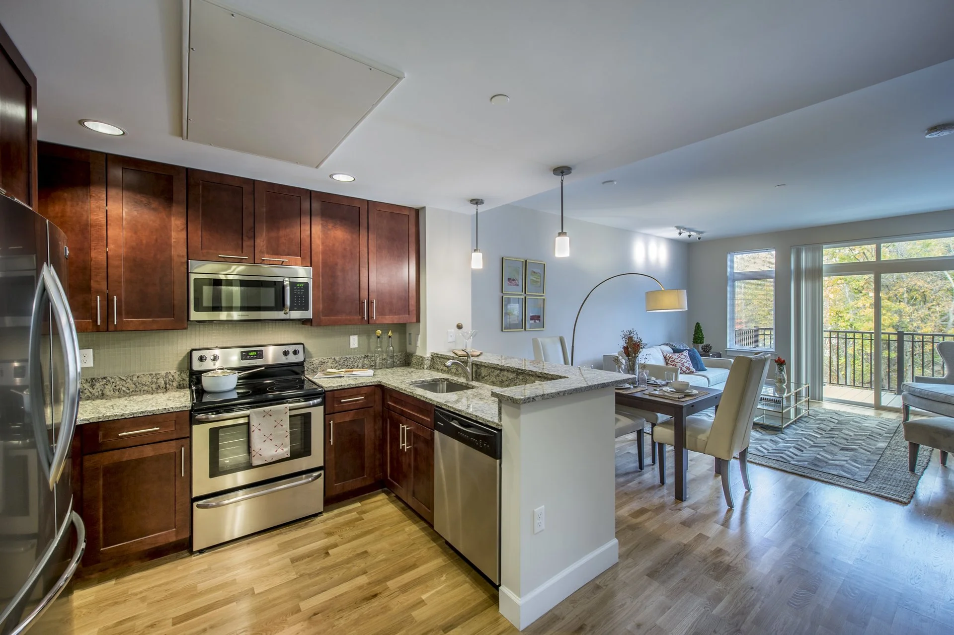 Open-concept kitchen and living area with wooden cabinets, stainless steel appliances, granite countertops, and a balcony with view of trees.