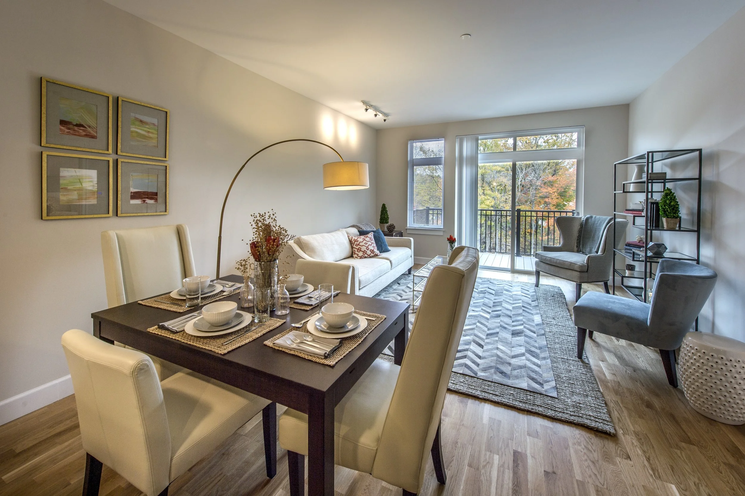 Living and dining room with a dining table set for four, a white sofa, multiple armchairs, a striped area rug, and a large window with a balcony view of trees.