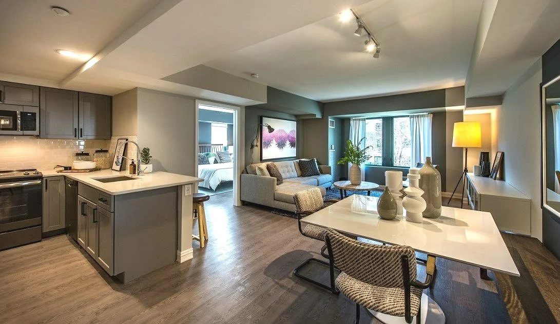 Open-concept living room and kitchen in a modern apartment with a dining table, sofa, and bedroom visible in the background.