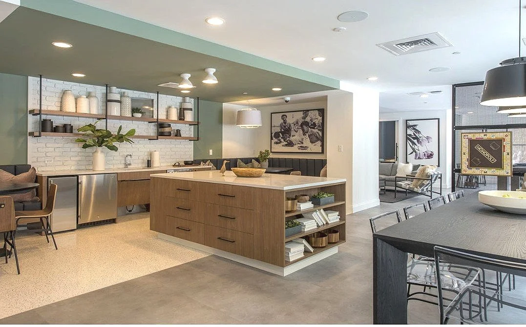 Modern open-concept kitchen and dining area with a kitchen island, open shelving, and a dining table with chairs. In the background, there is a sitting area with couches and framed artwork.