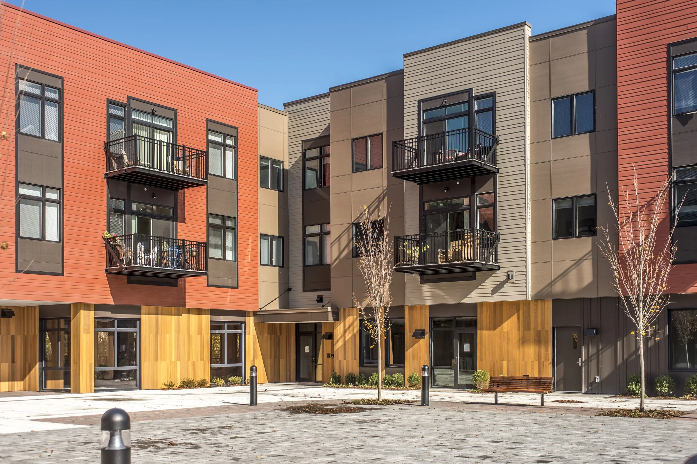 Modern apartment building with colorful exterior, small balconies, and landscaped courtyard with trees and benches.