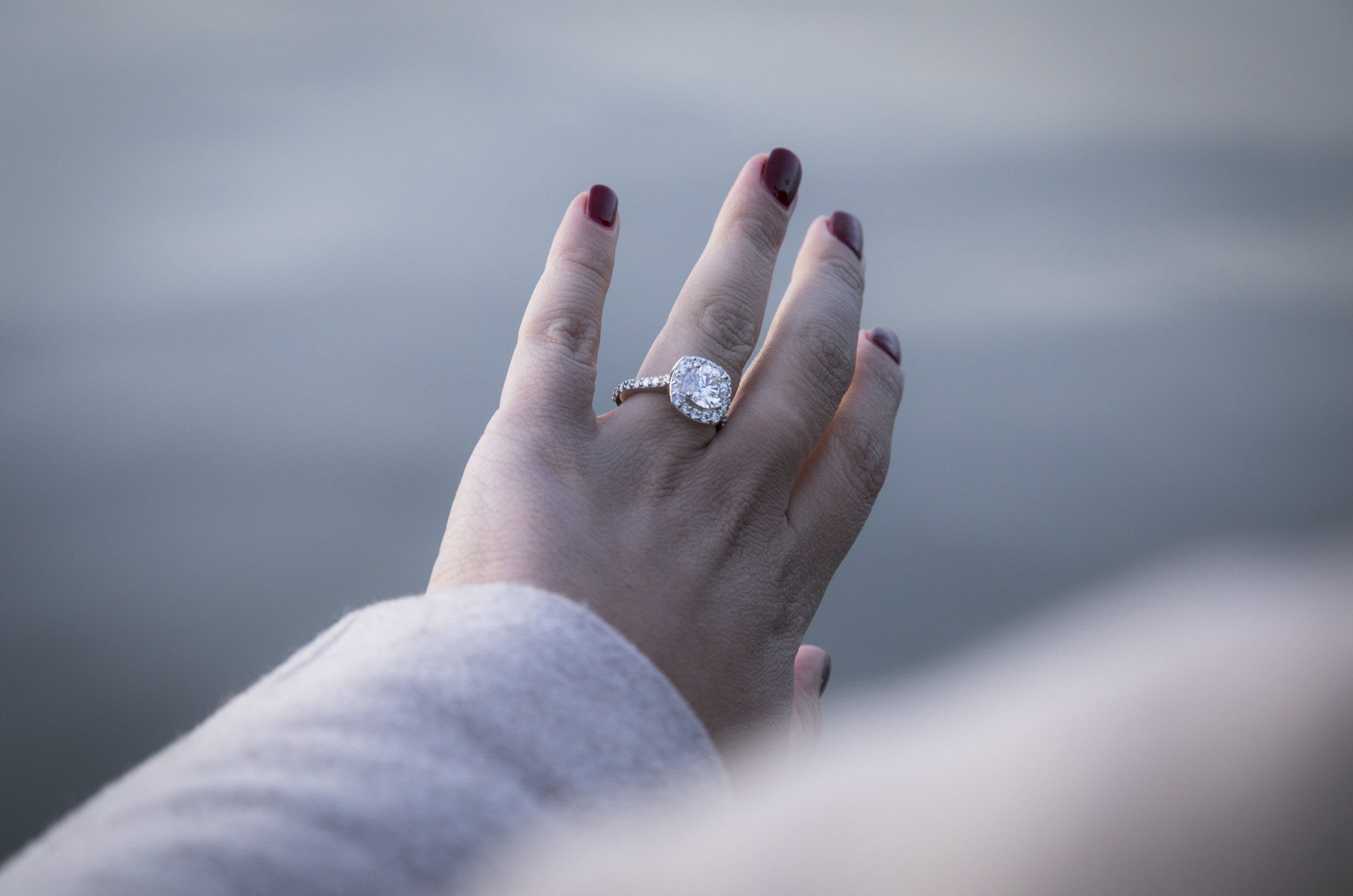 Close-up of a woman's hand with dark red nail polish, showcasing a large engagement ring with a cushion-cut diamond and a band of smaller diamonds, against a blurred outdoor background.