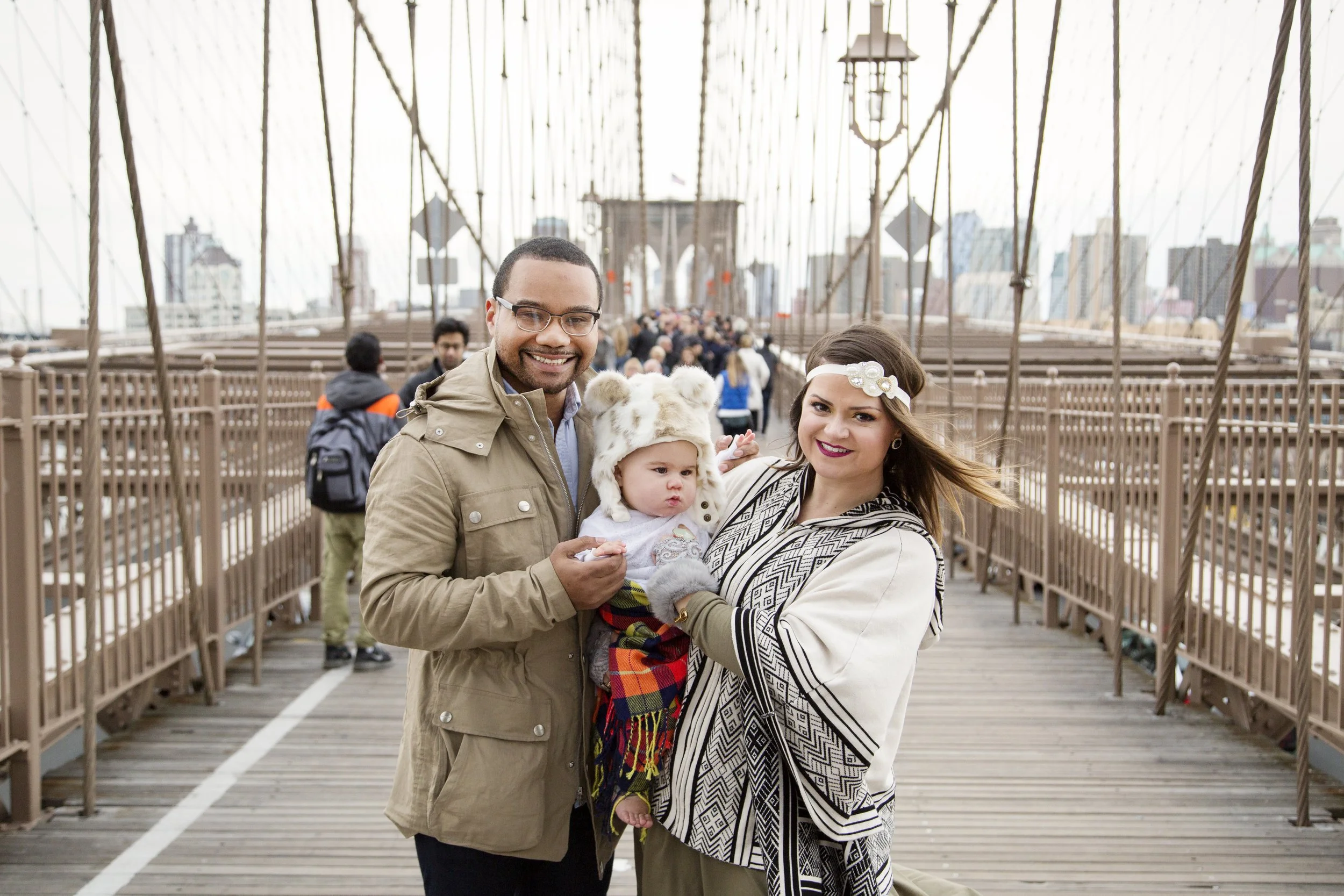 A family of three standing on the Brooklyn Bridge in New York City, smiling at the camera, with city buildings in the background.