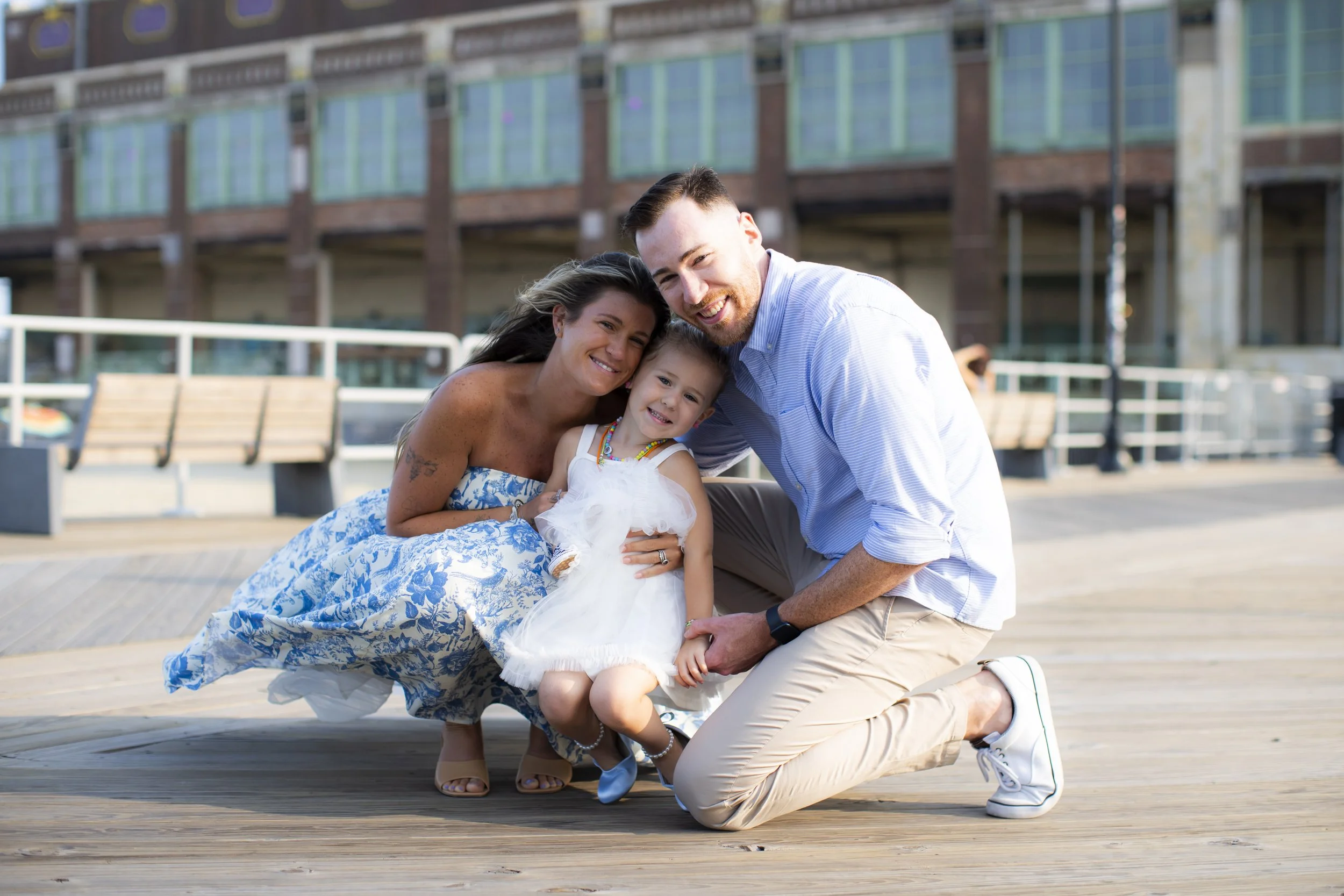 A happy family of three smiling and kneeling on a wooden dock outside in front of a modern building, with a woman in a blue and white patterned dress, a man in a blue shirt and beige pants, and a young girl in a white dress.