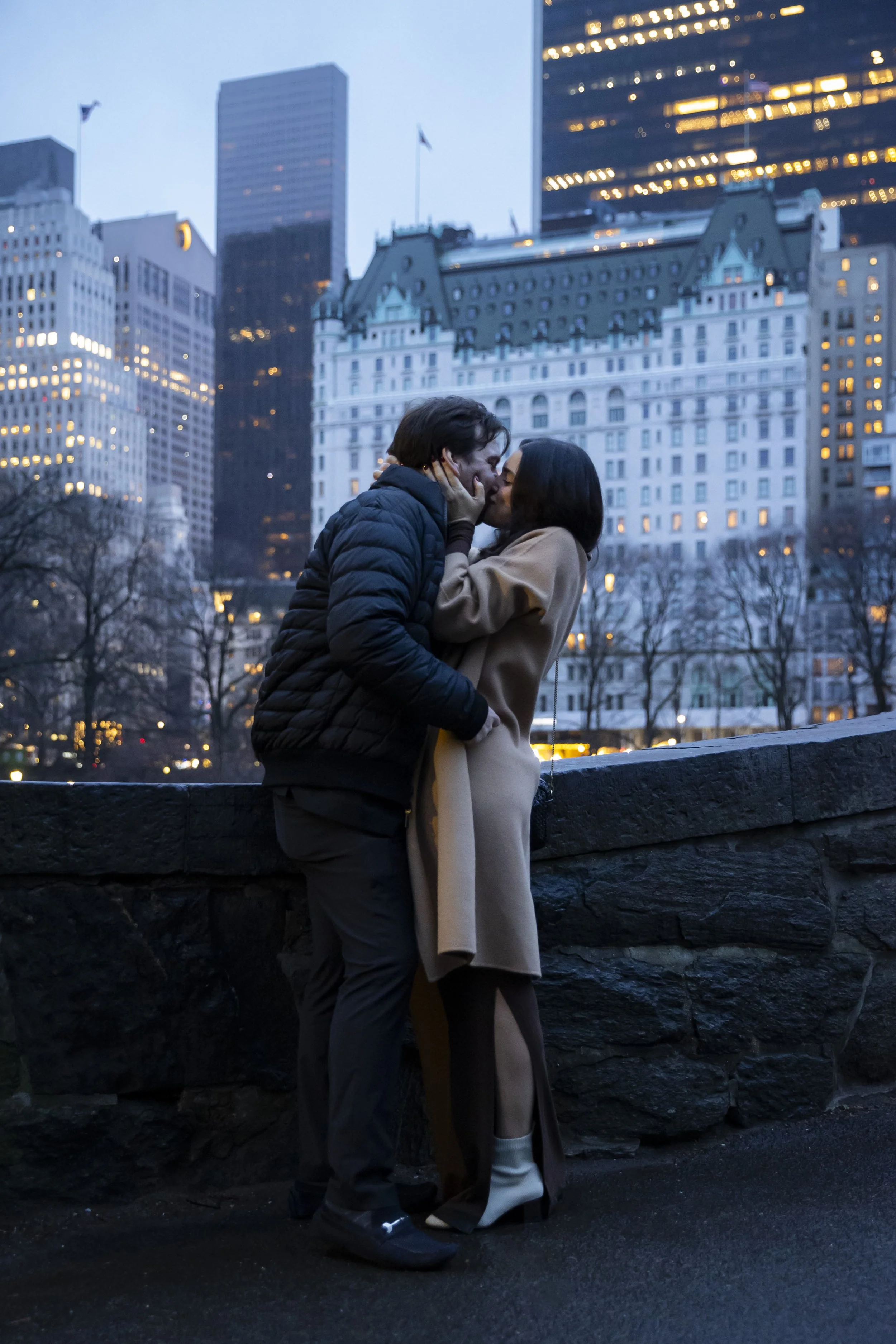 A couple sharing a kiss on a city sidewalk at dusk, with tall buildings and city lights in the background.