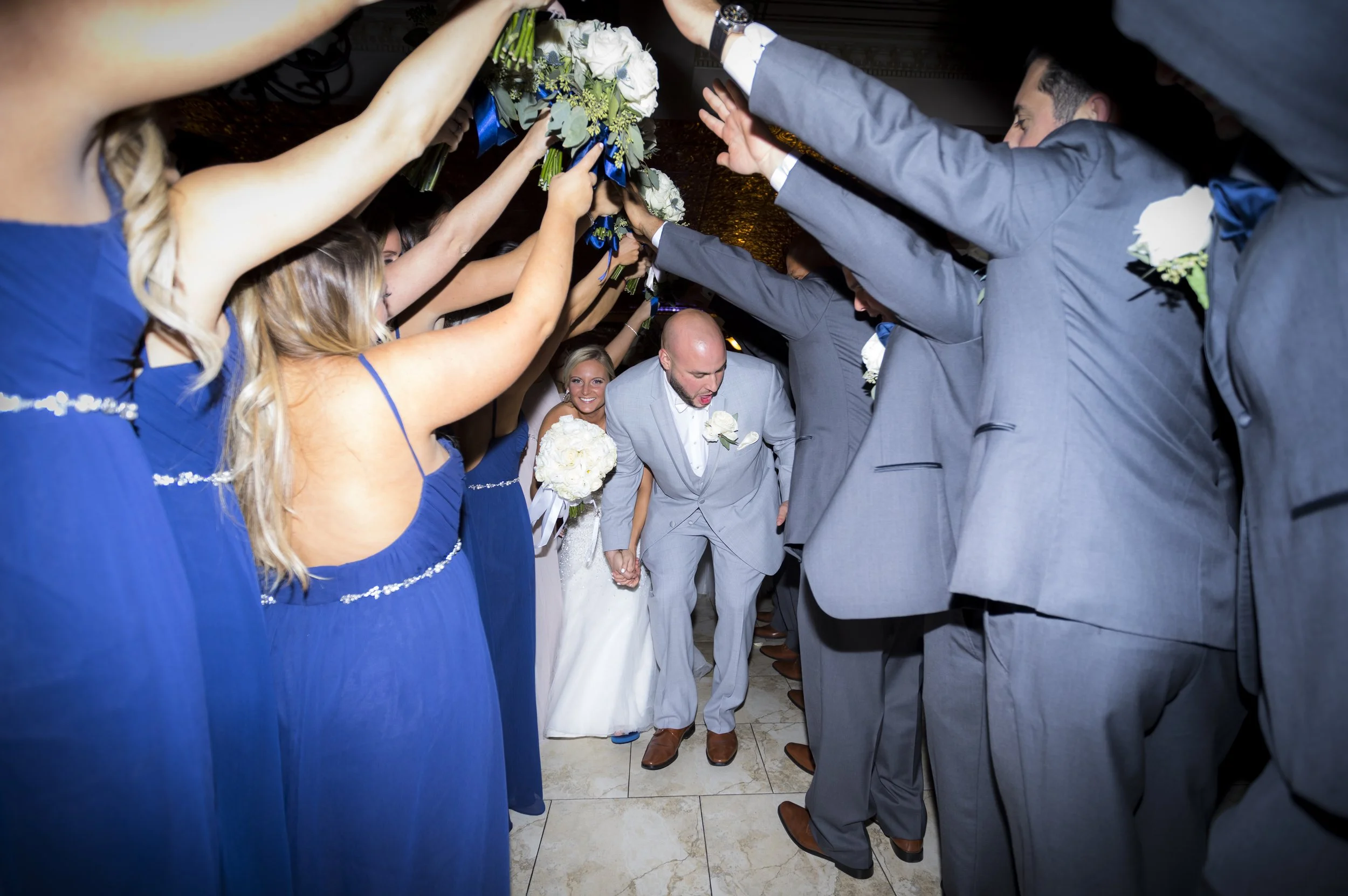 Bride and groom walking through a tunnel of bridesmaids and groomsmen holding bouquets and wearing matching blue and gray suits
