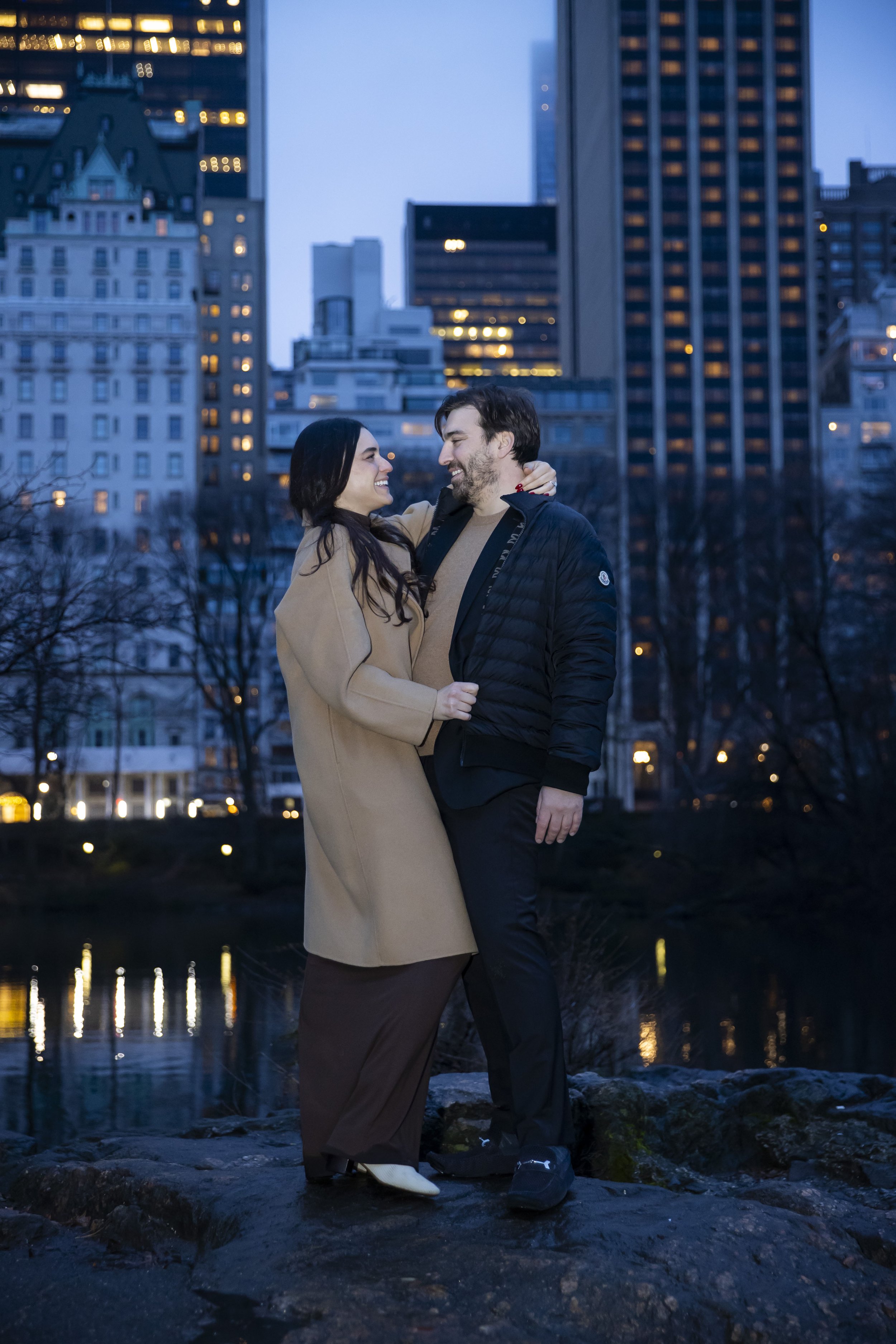 A couple standing on a rocky surface by a river in an urban park at dusk. They are smiling and looking at each other, with the city skyline and tall buildings lit up in the background.