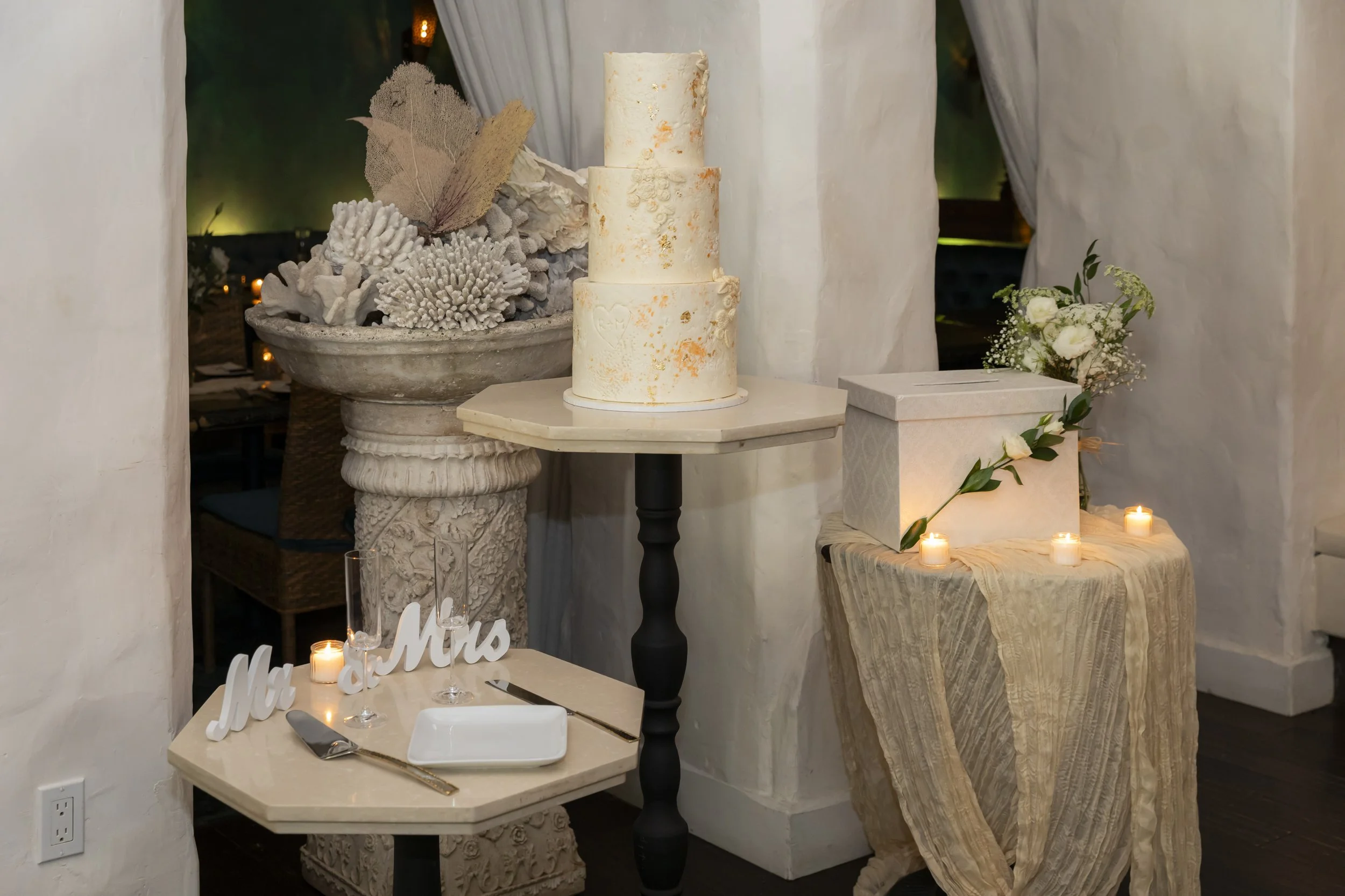 Decorative wedding setting with a three-tiered white cake on a black stand, candles, floral arrangements, Mr. and Mrs. signs, and textured fabric draped over tables.