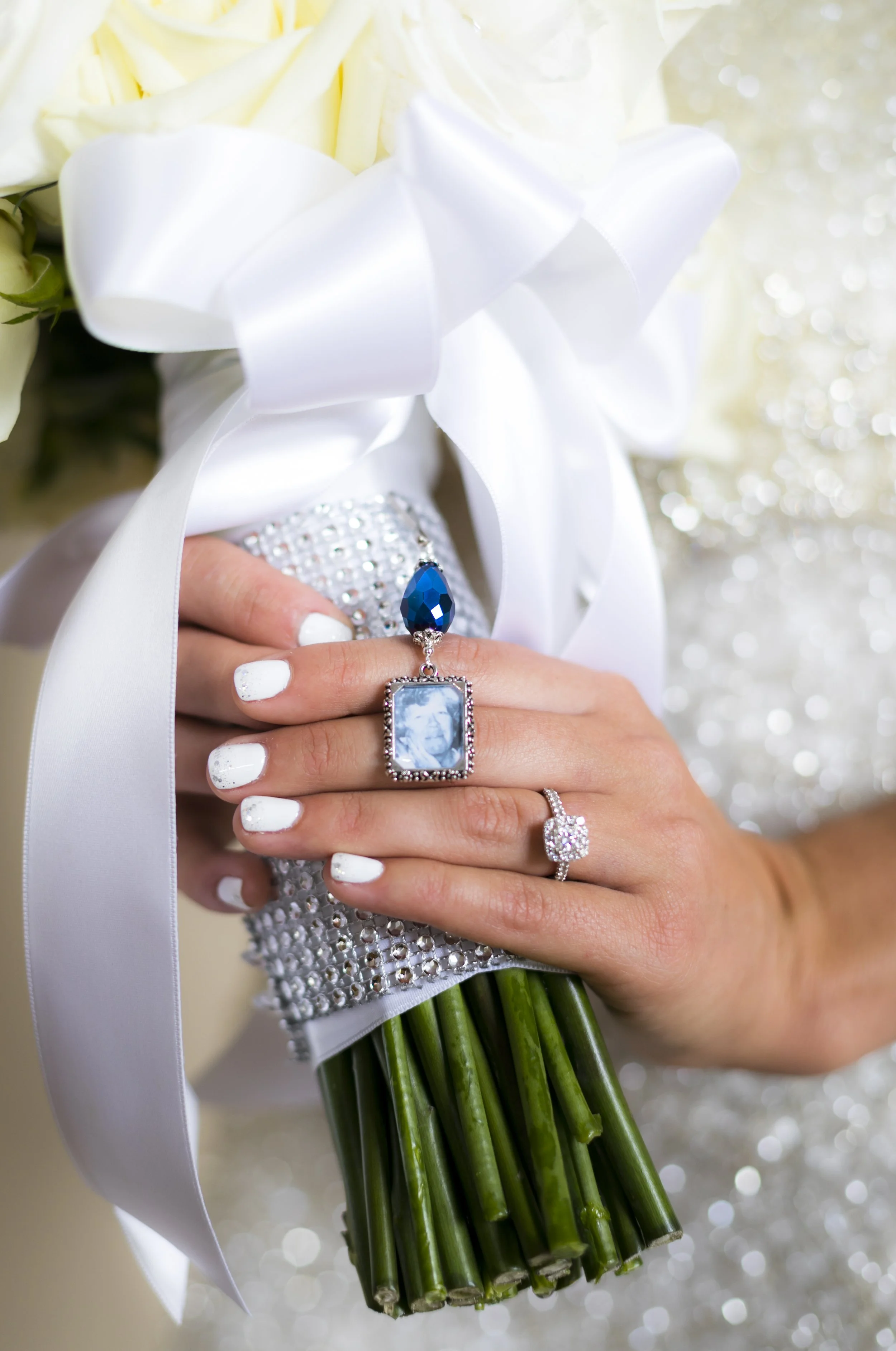 A bride holding a wedding bouquet with white roses, adorned with a large satin bow and diamond accents. The bride's hand displays an elaborate engagement ring and a wedding band, with a blue gemstone pendant hanging from her ring finger. The bouquet 
