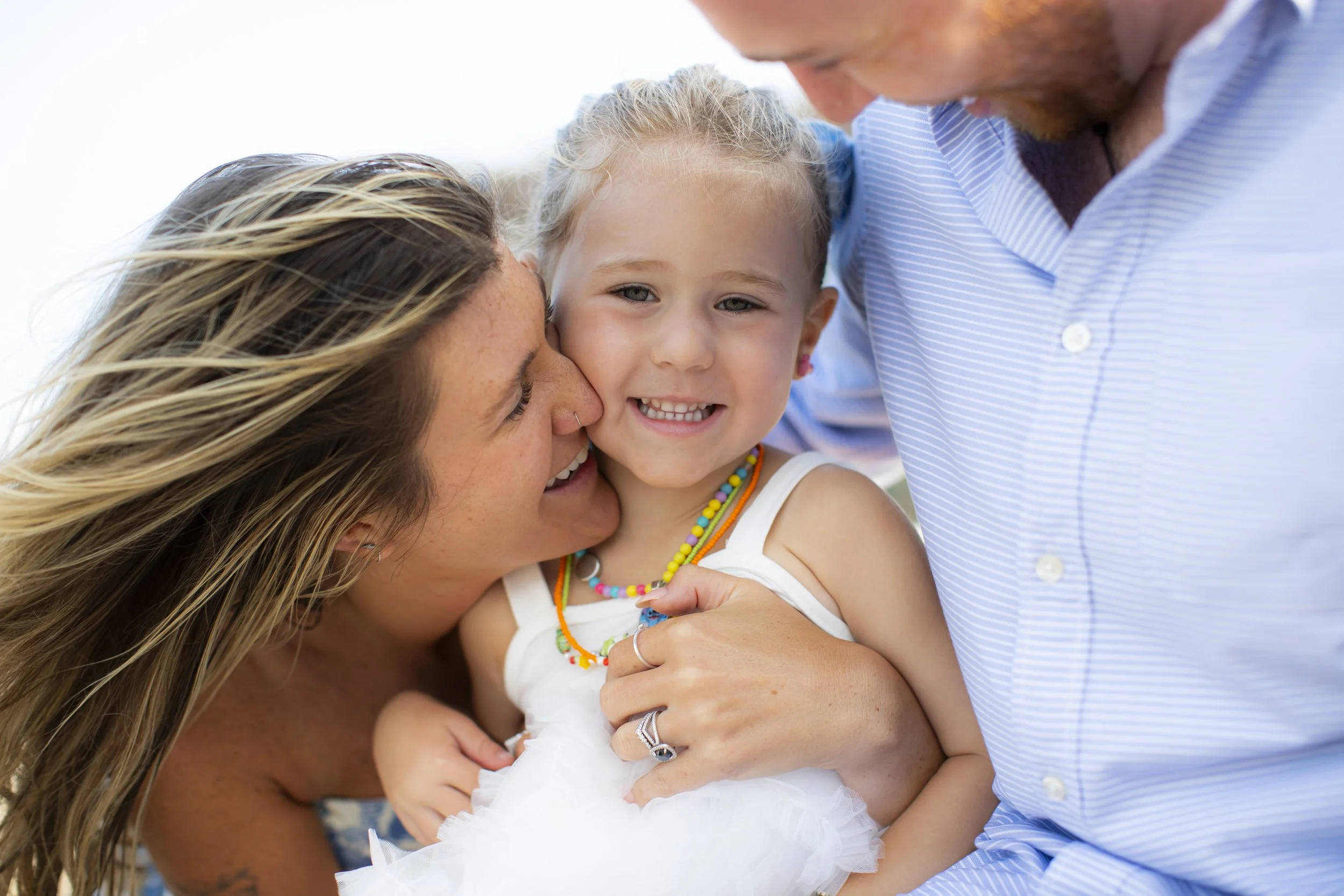 Family of three, woman, man, and young girl, smiling and embracing outdoors on a sunny day.