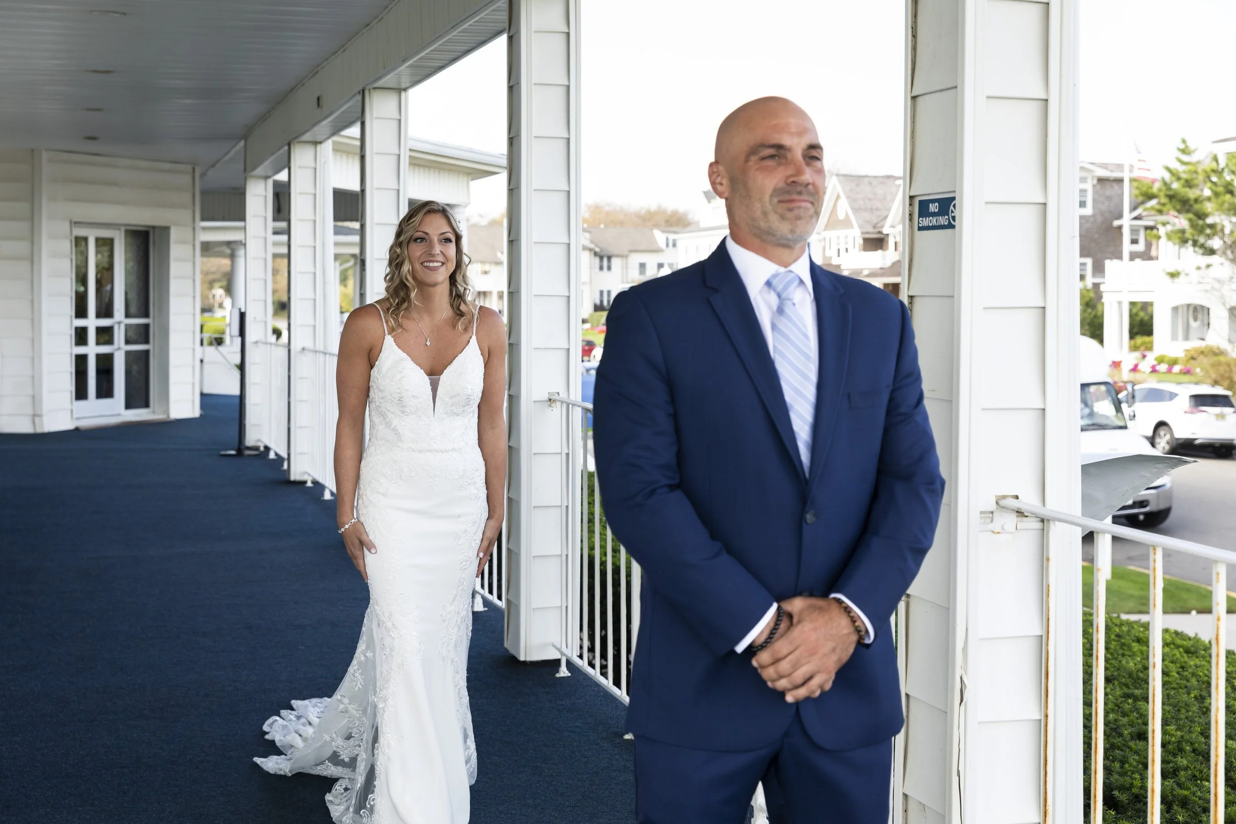A bride in a white wedding dress looks at a man in a blue suit who appears to be nervous and awaiting her. They are standing on a balcony outside a white building with parking visible in the background.