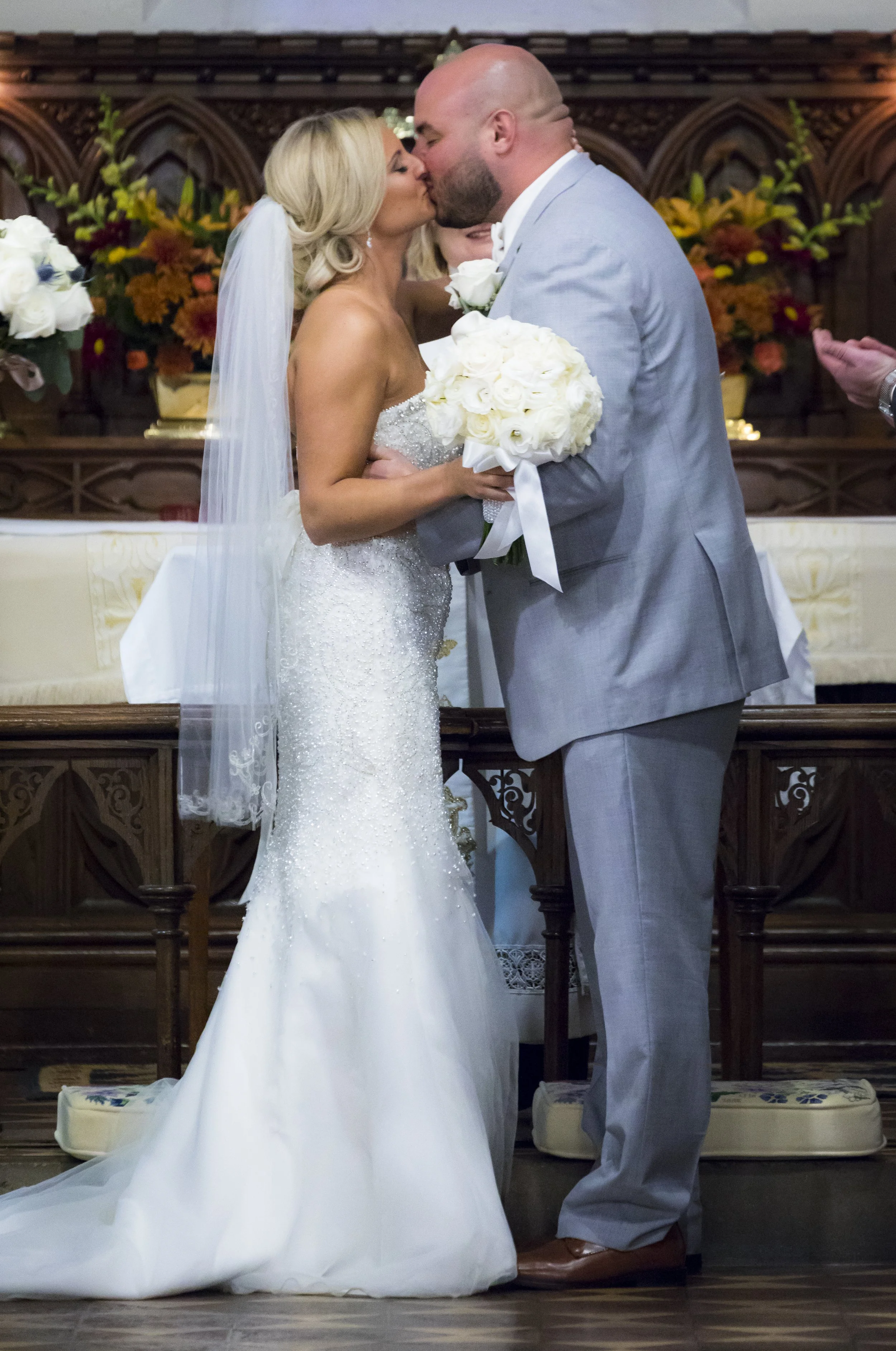 A bride and groom share a kiss during their wedding ceremony, with the bride holding a bouquet of white roses and wearing a strapless white wedding gown. The groom is dressed in a light gray suit.