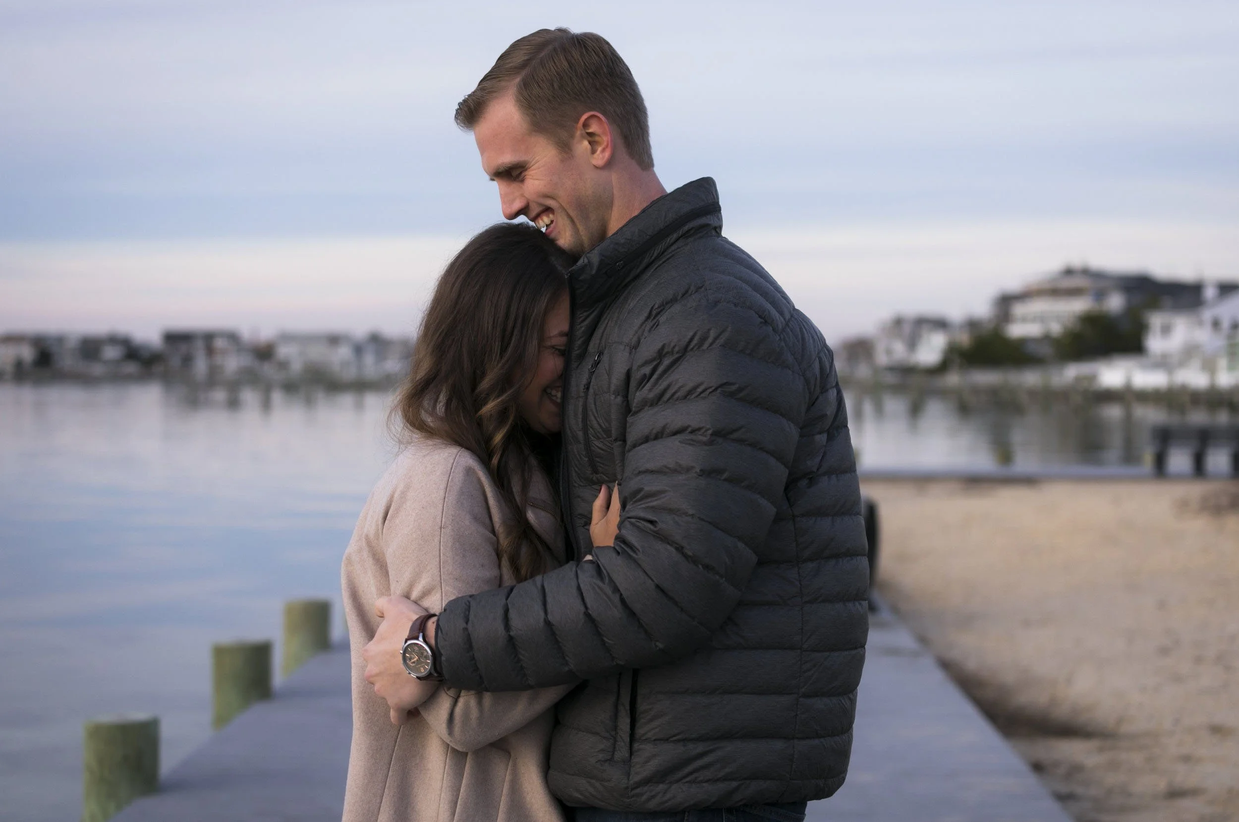 A couple hugging on a dock beside a calm body of water at sunset, with houses and boats in the background.