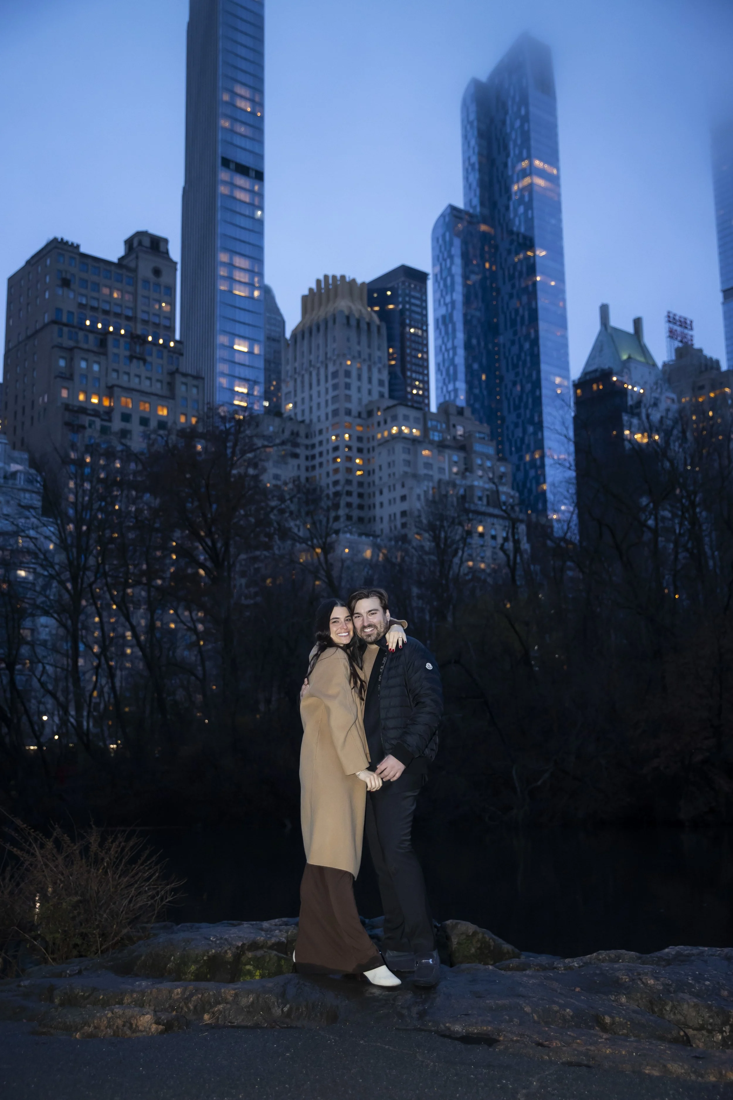 A smiling couple standing on rocks with a city skyline at dusk in the background.