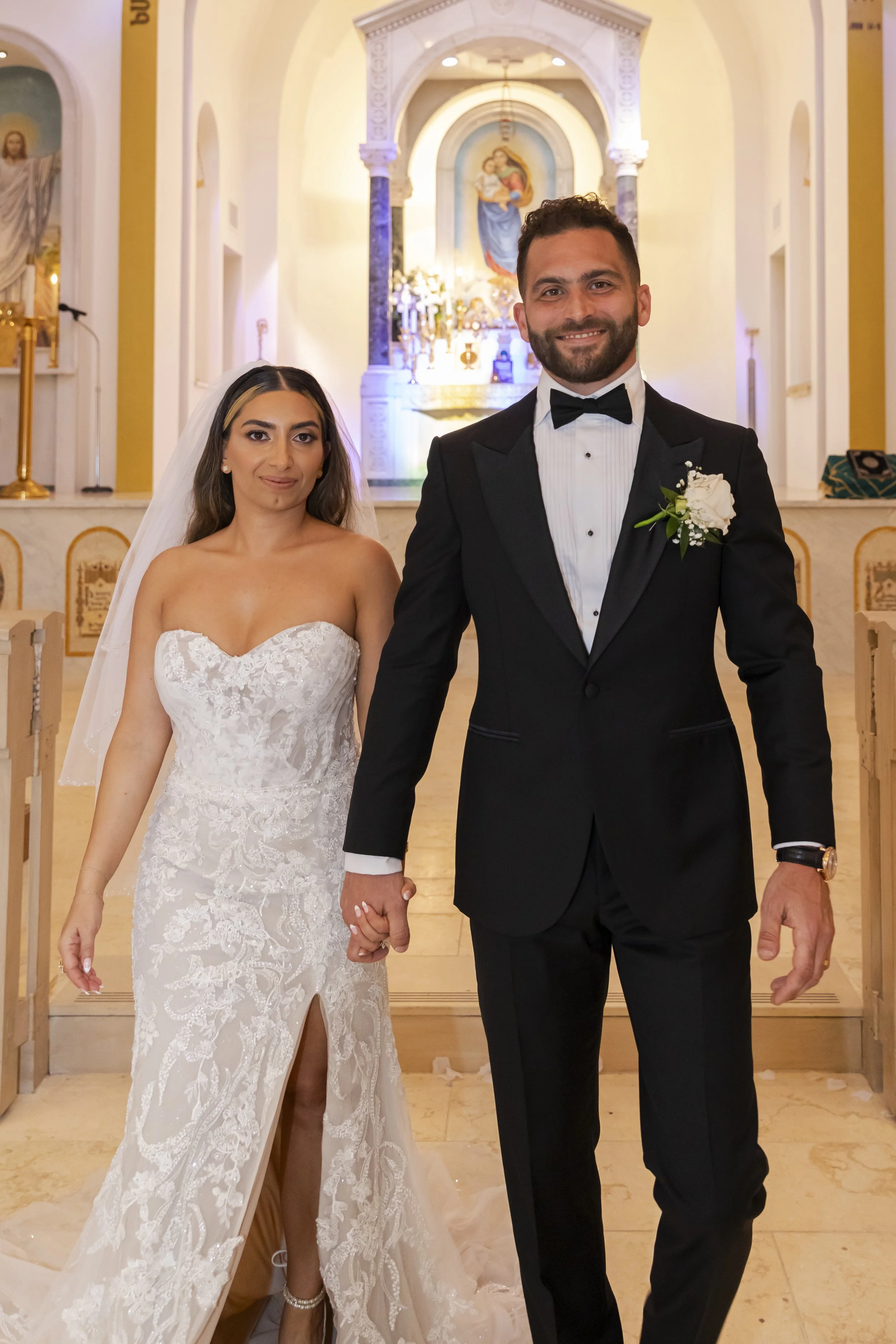 A bride and groom holding hands inside a church, with an altar and religious artwork in the background.