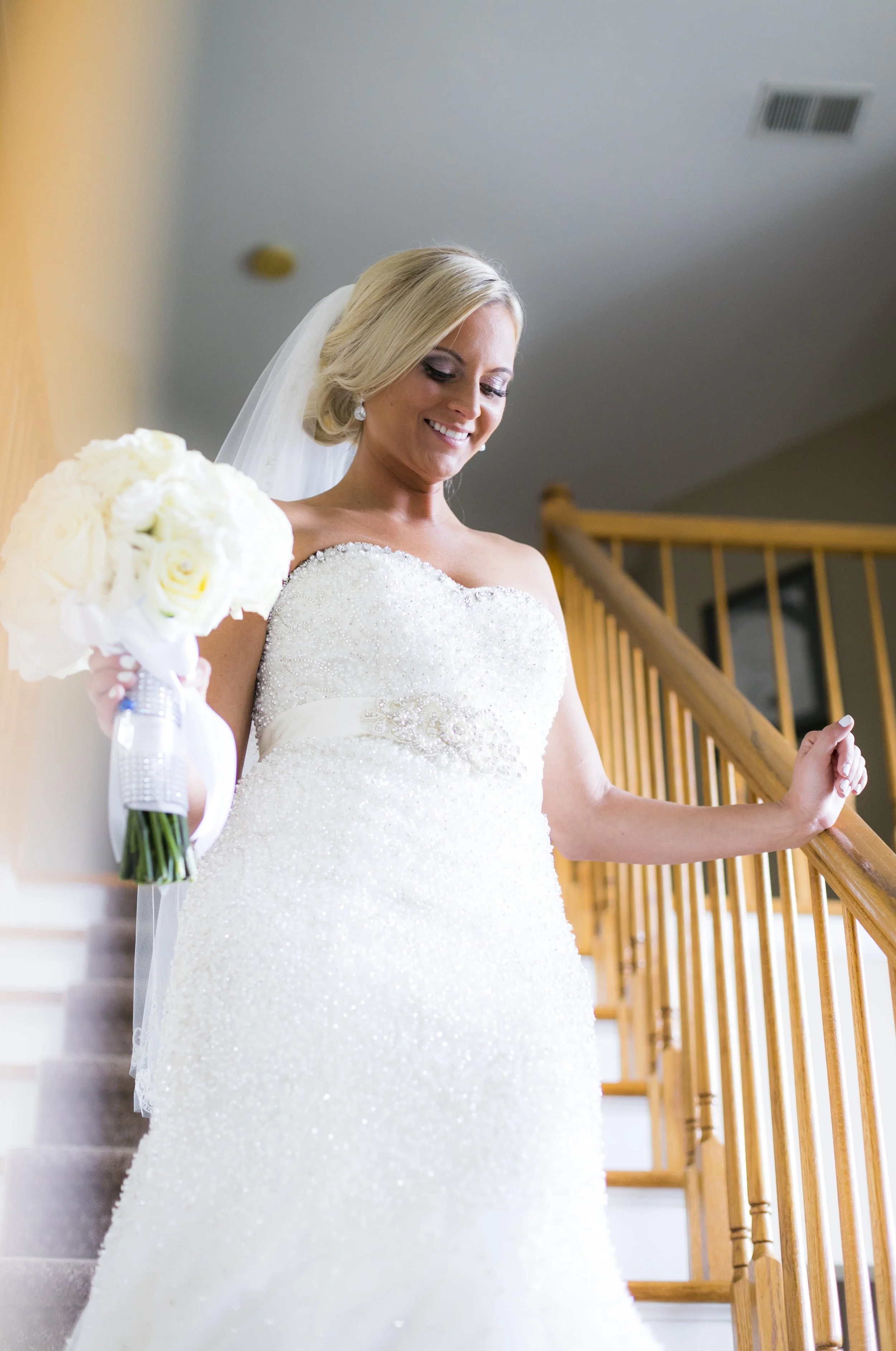 Bride in a white wedding gown holding a bouquet of white roses, standing on stairs and smiling.