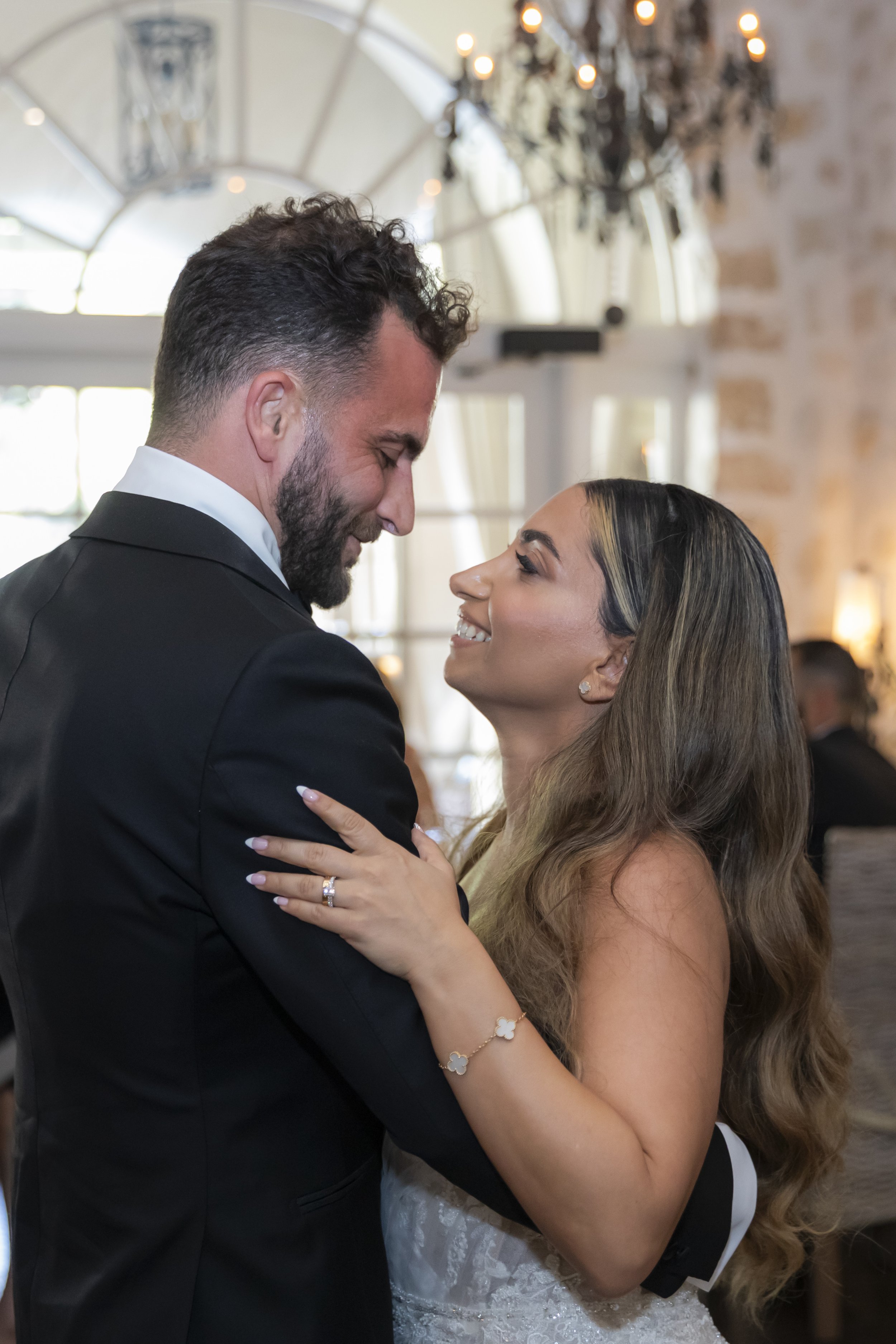 A man and woman dressed in formal wedding attire sharing a dance, smiling at each other in a beautifully lit indoor setting.