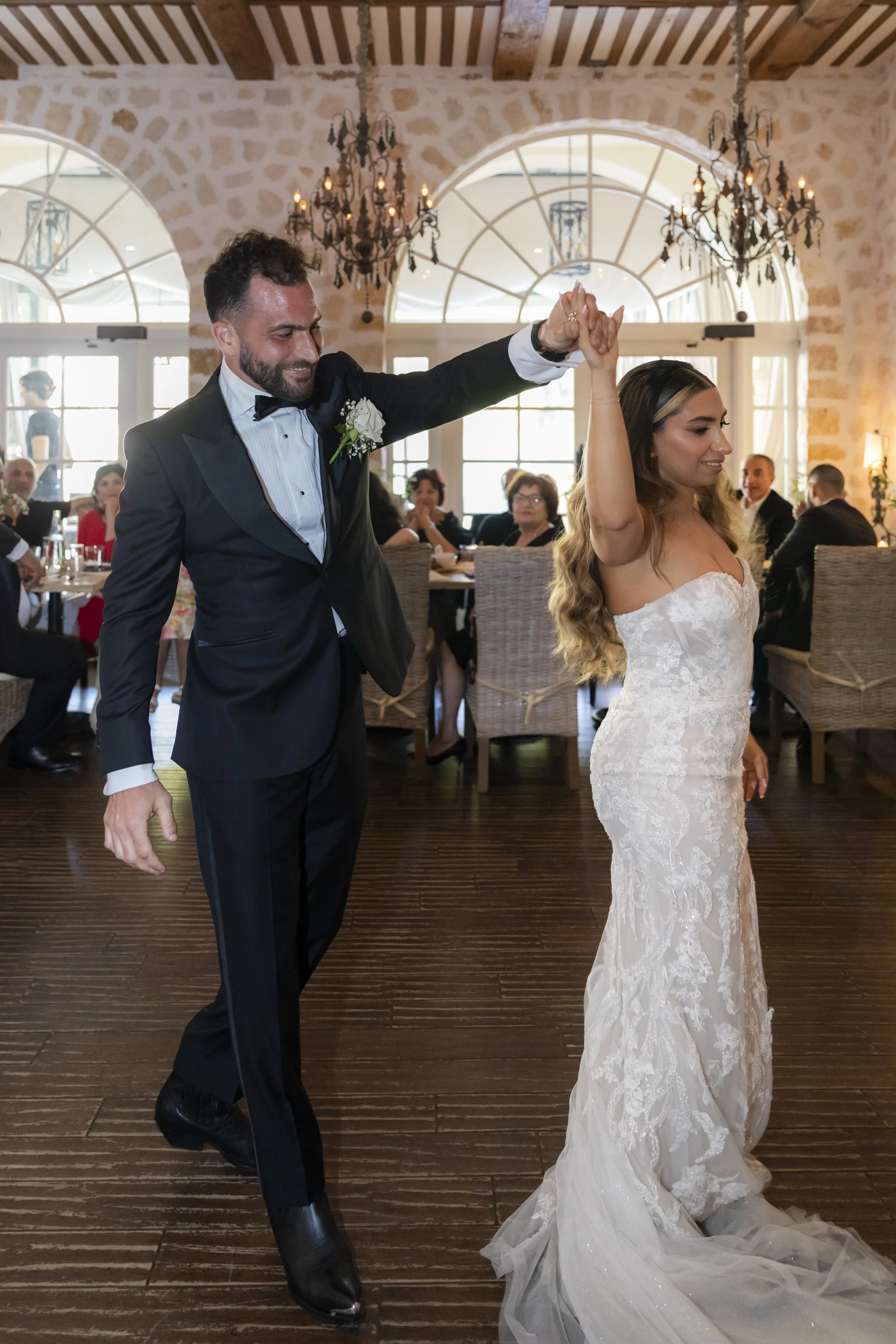 A newly married couple dances at their wedding reception inside a rustic venue with stone walls, large windows, and chandeliers. The groom wears a black tuxedo with a white shirt and bow tie. The bride wears a strapless white lace wedding gown with l
