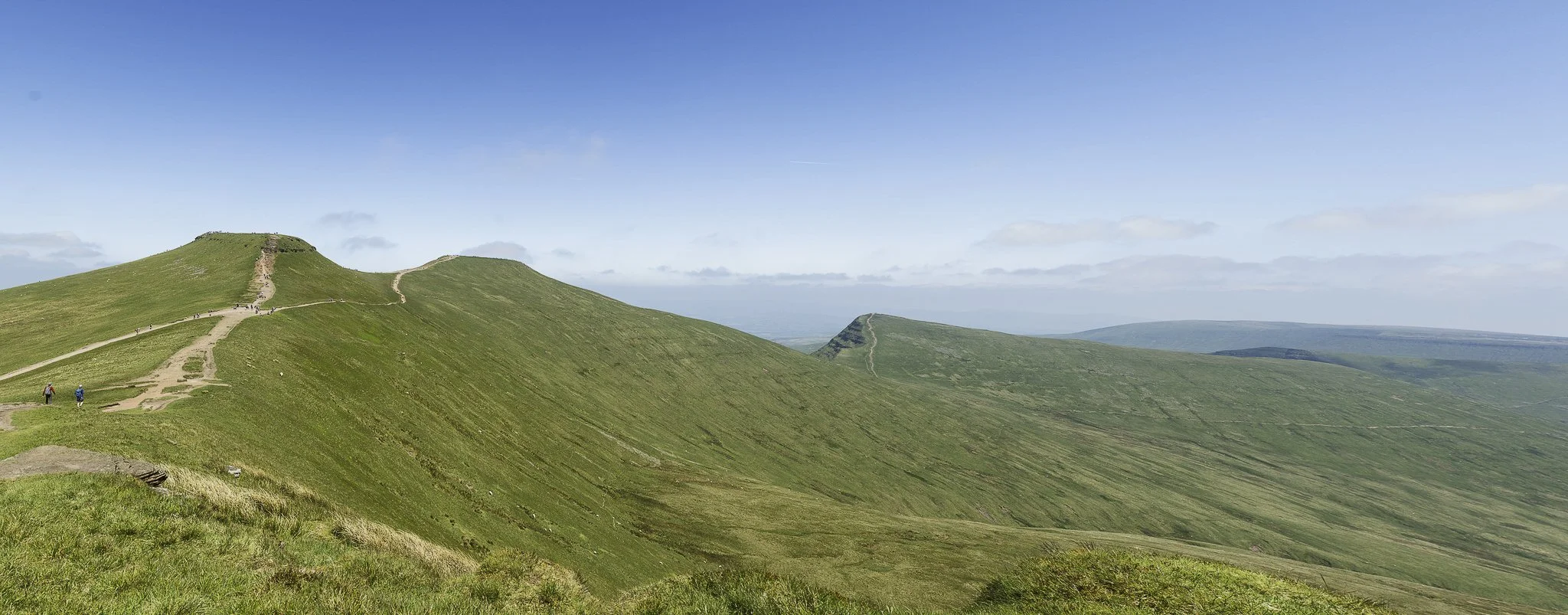 Green rolling hills with narrow dirt paths, some hikers walking, under a blue sky with scattered clouds.