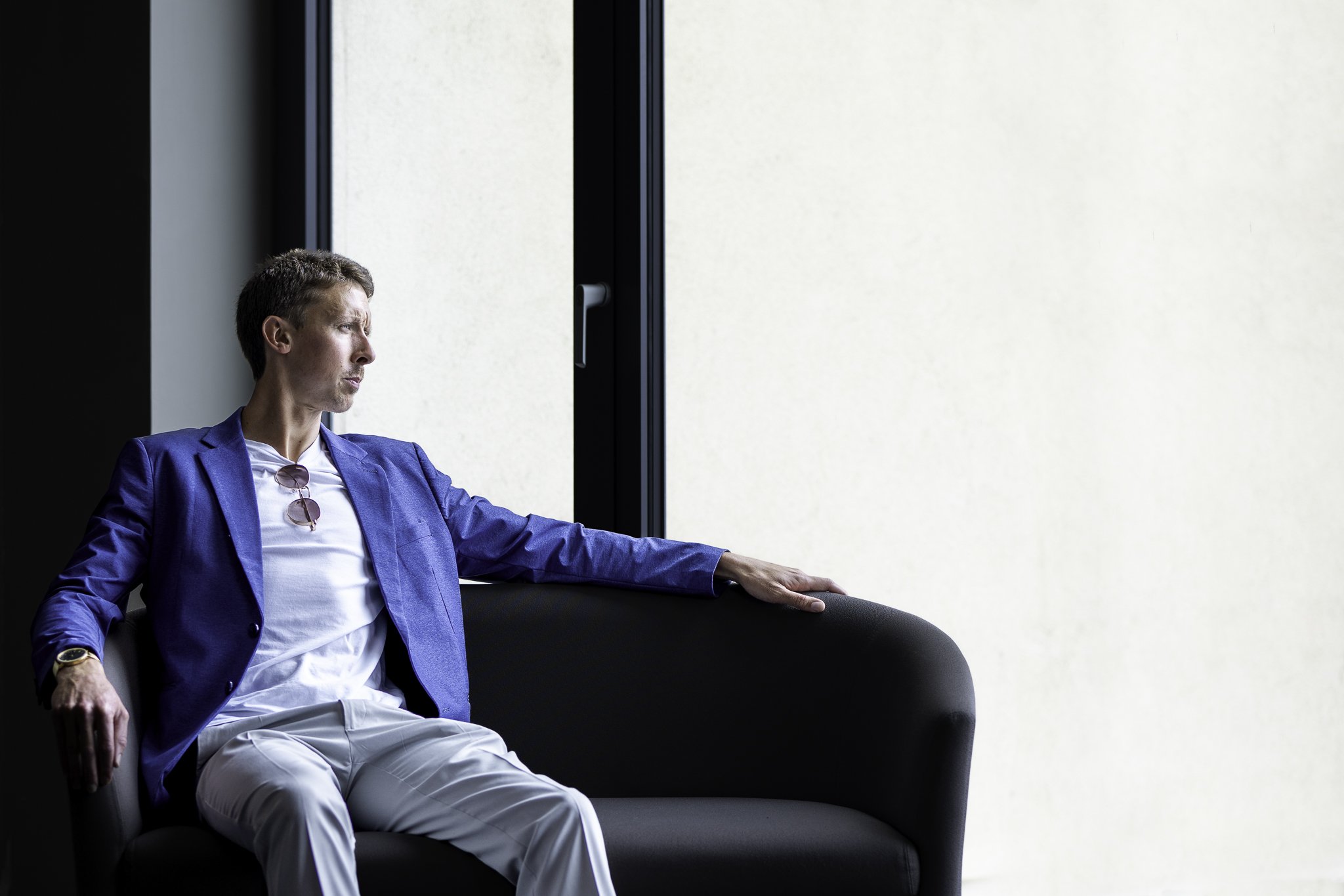 Man in a blue blazer and white pants sitting on a black sofa by a window, looking outside.