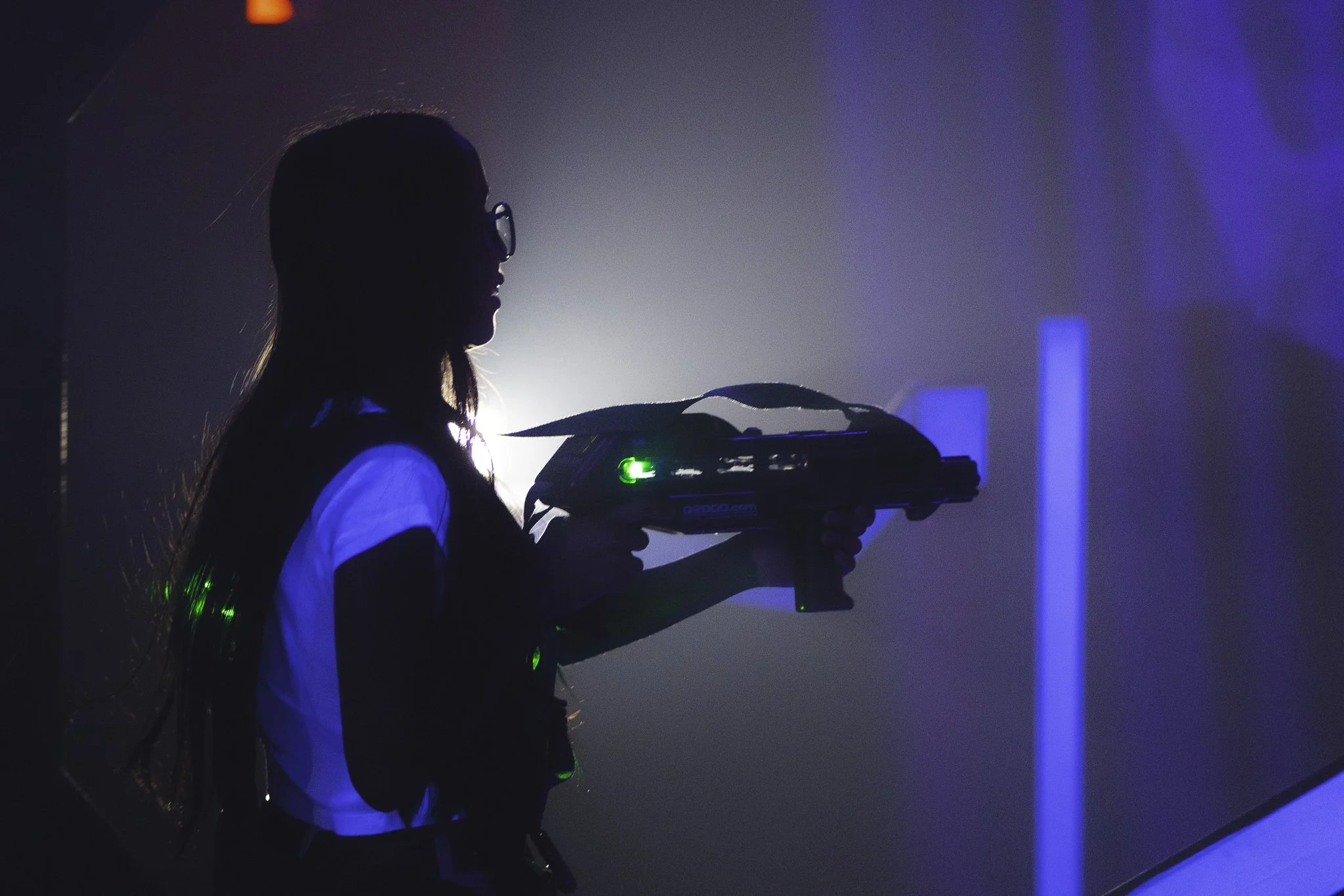 Silhouette of a woman wearing glasses holding a laser tag gun in a dark room illuminated by blue and green lights.
