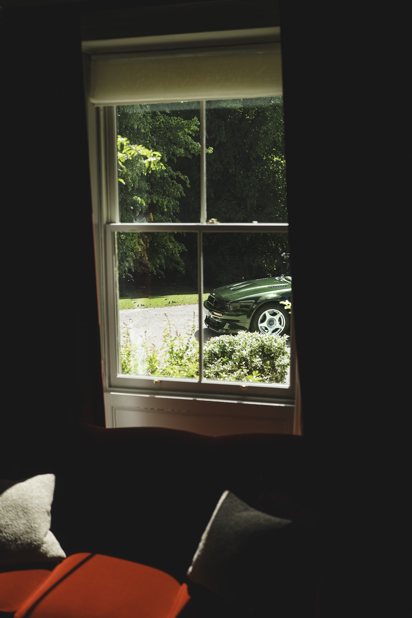 Looking through a window, a black car is parked outside on a gravel driveway, surrounded by green trees and bushes.