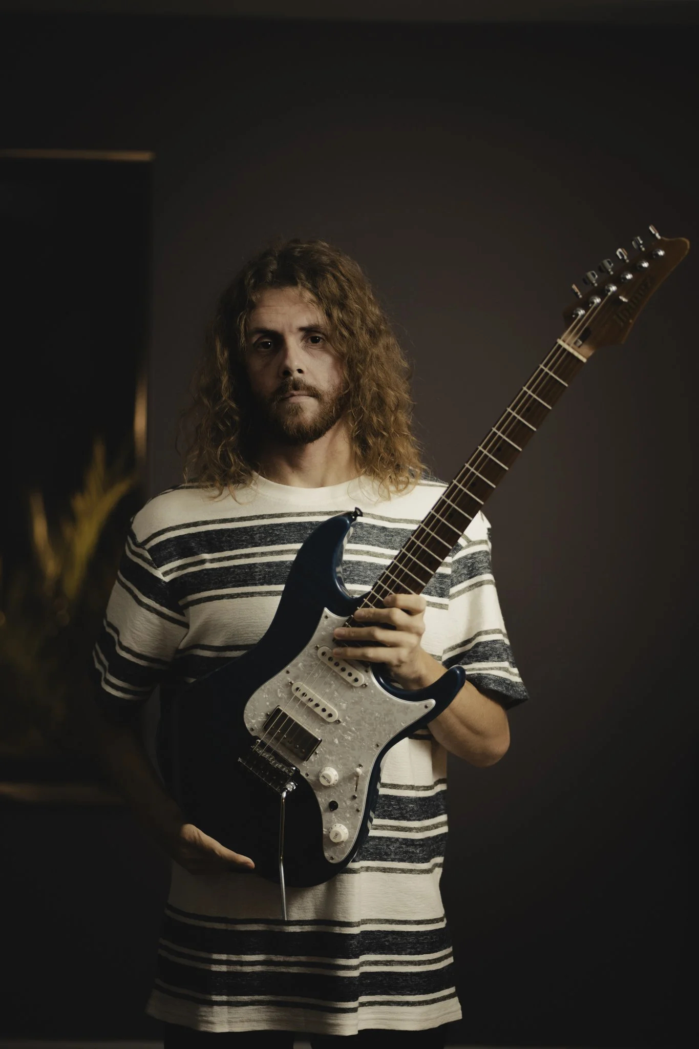 A man with long curly hair and a beard holding an electric guitar standing against a plain dark background.