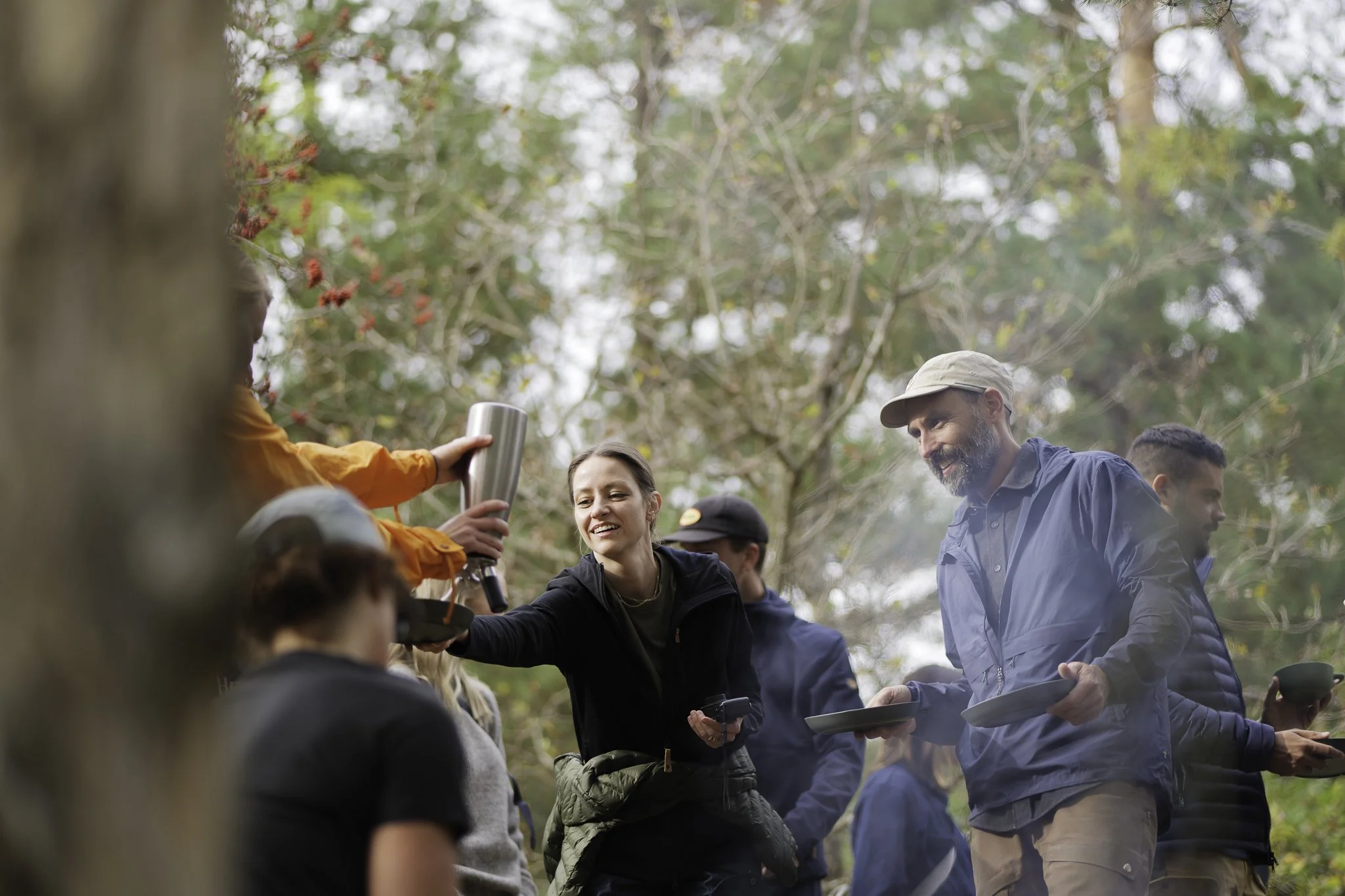 People gathering outdoors in a forest, pouring drinks, and socializing.