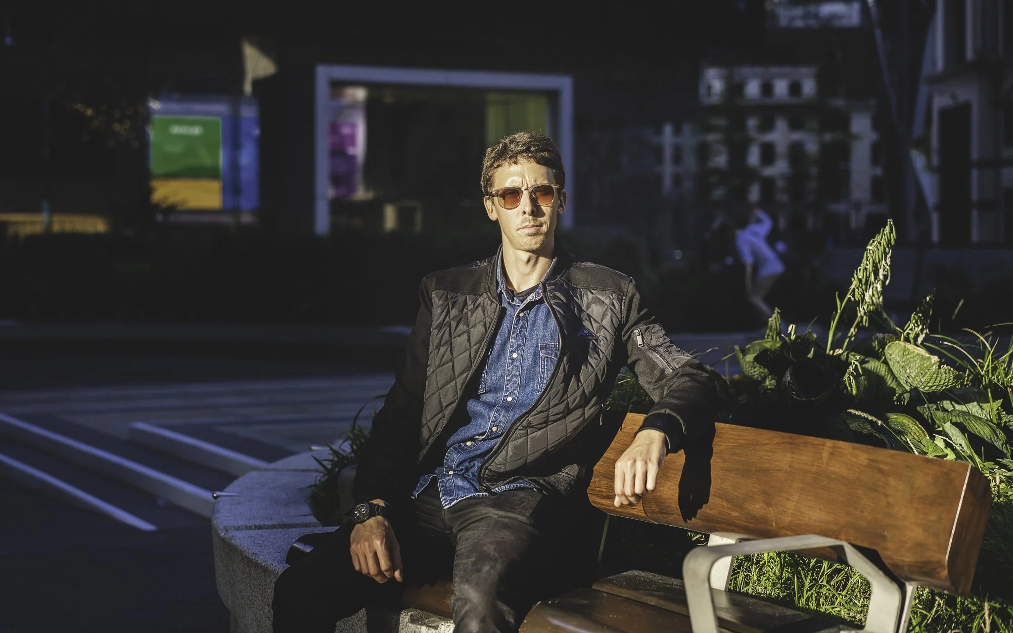 A young man with short brown hair, wearing sunglasses, a black quilted jacket, a blue denim shirt, and black pants, sitting on a wooden park bench during daytime with sunlight highlighting his face. In the background, there are some buildings and a b