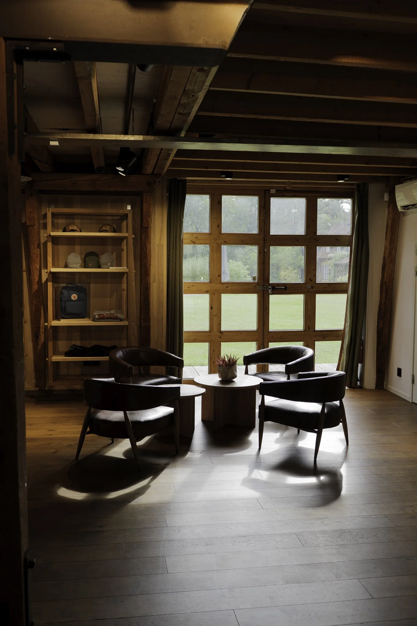 A cozy indoor seating area with three dark leather chairs arranged around a central round wooden table, a potted plant on top, large wooden-framed windows with green curtains, wooden walls, and a view of a grassy yard outside.