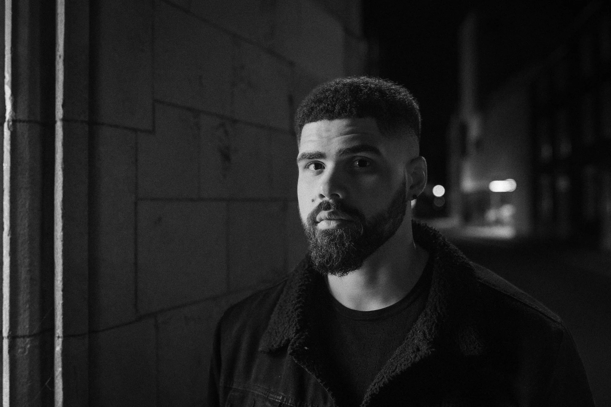 A young man with a beard and short hair standing outdoors at night near a brick wall, illuminated by streetlights, looking at the camera.