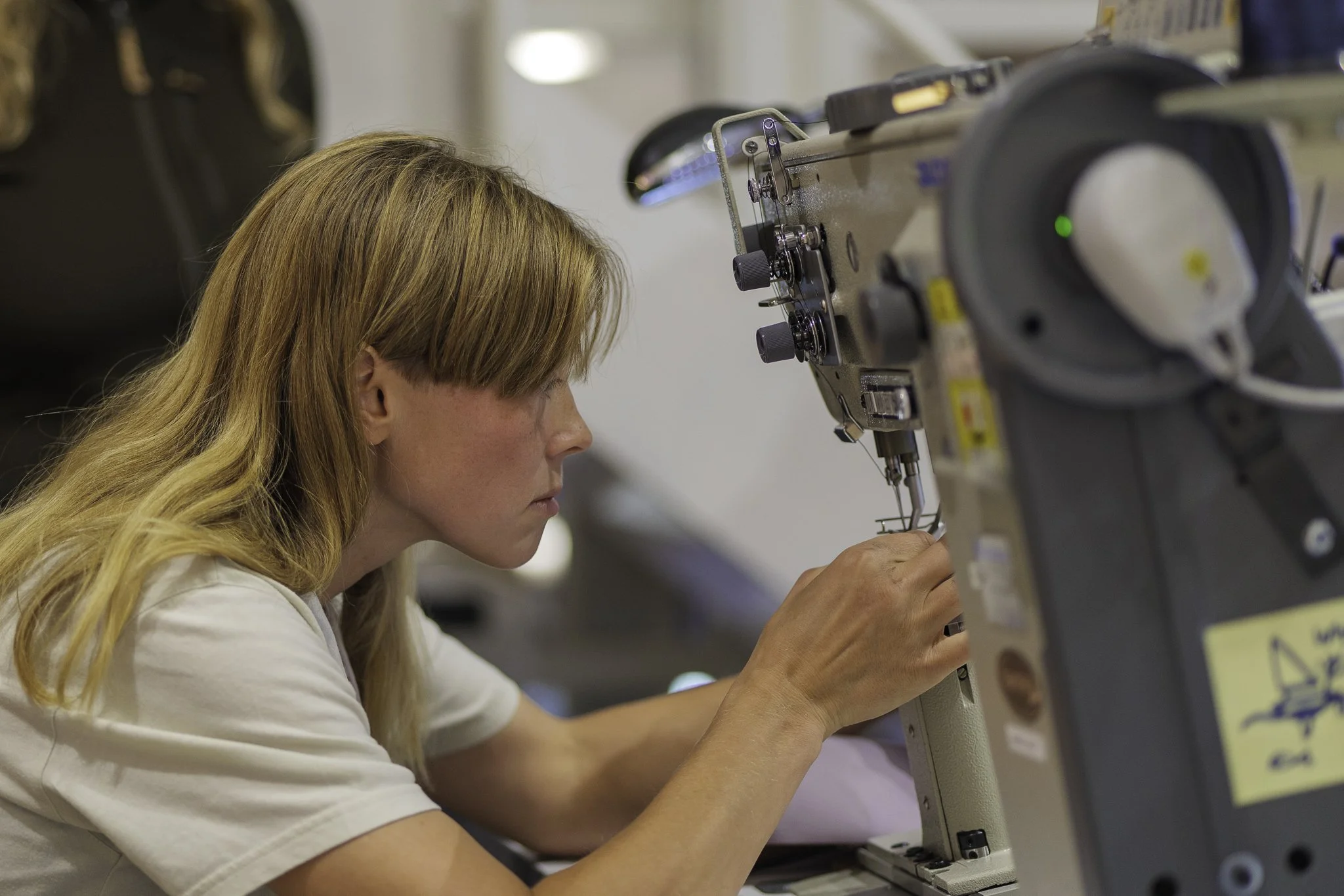 A woman with long blonde hair operating a sewing machine in a workshop or factory.