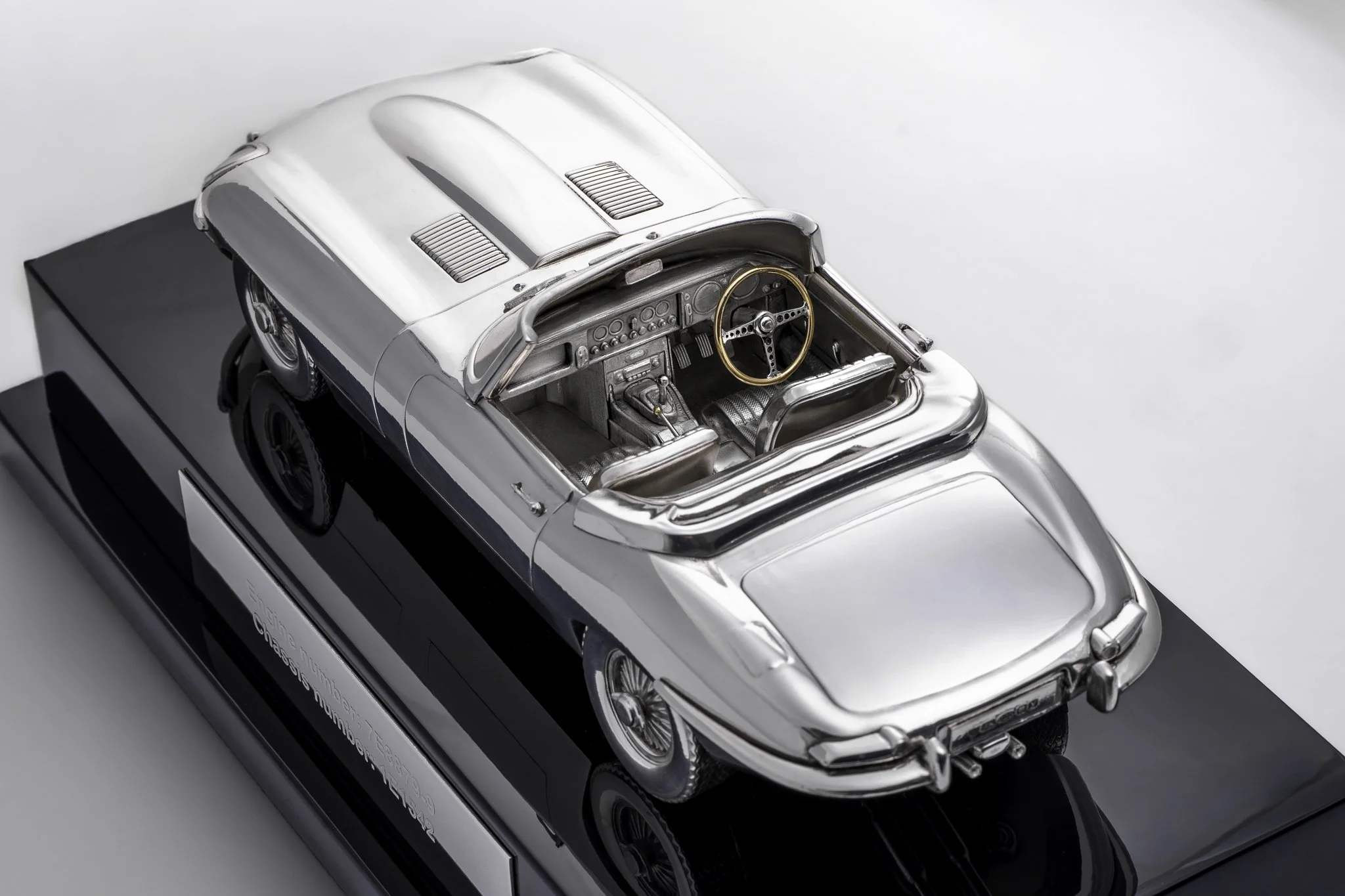 A silver model race car with an open interior showing the dashboard and steering wheel, displayed on a black stand with a nameplate.