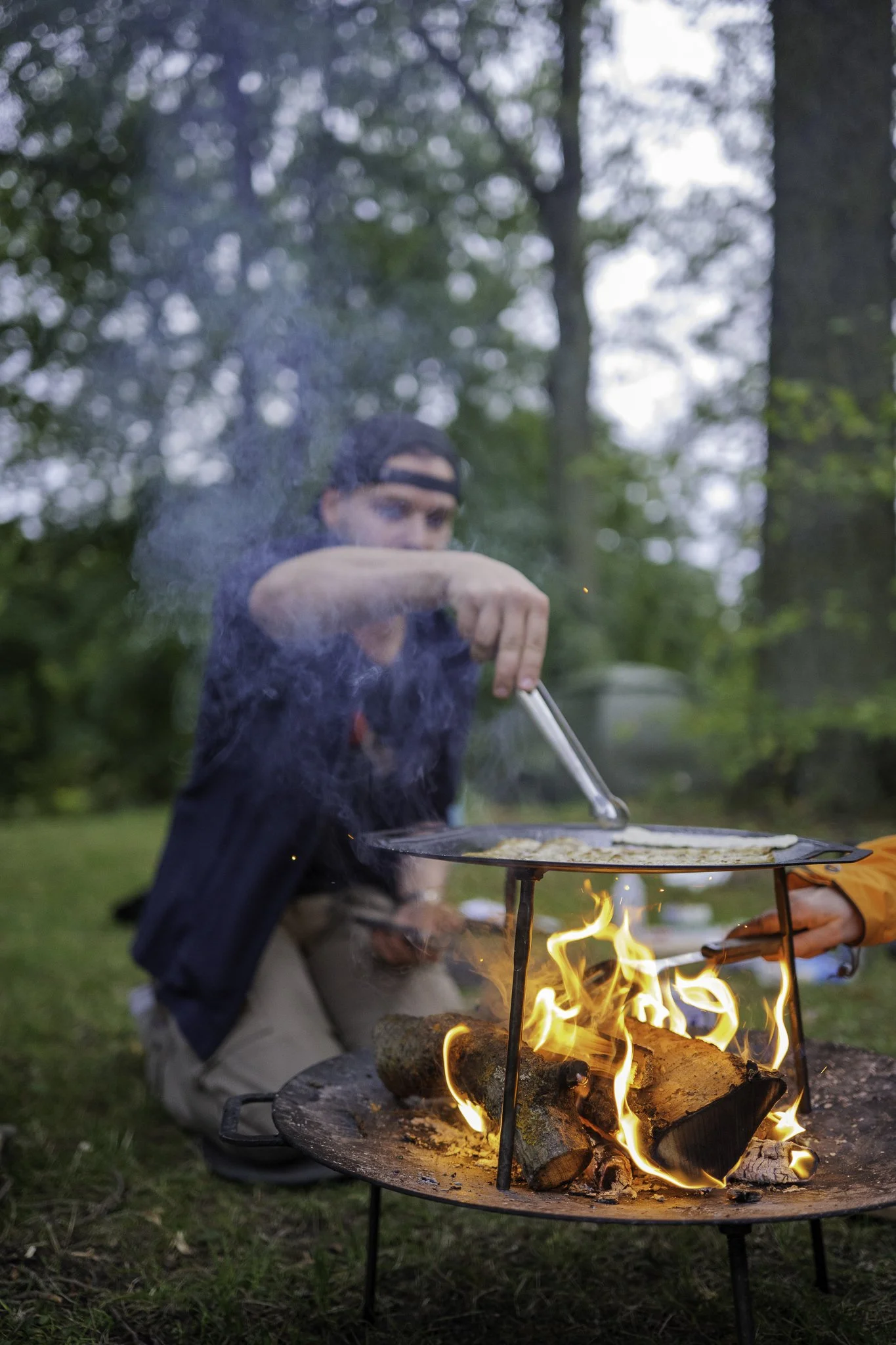 A person cooking food over an outdoor fire on a grill with wood logs, surrounded by trees in a forested area.