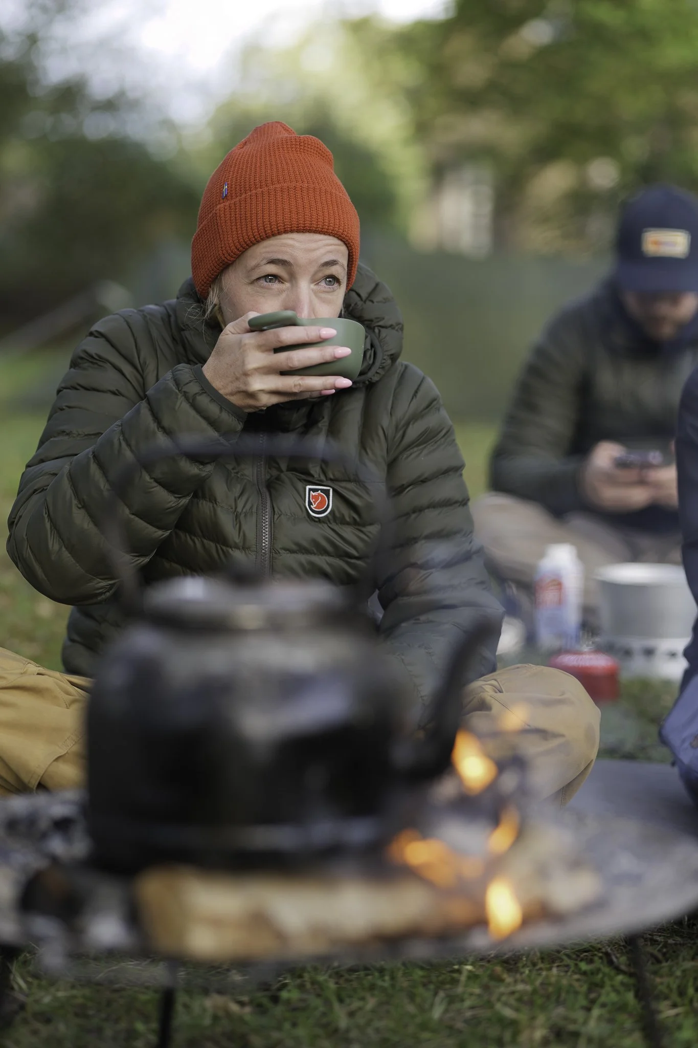 Person in a red beanie and green jacket drinking from a cup outdoors, with a campfire and another person in the background.